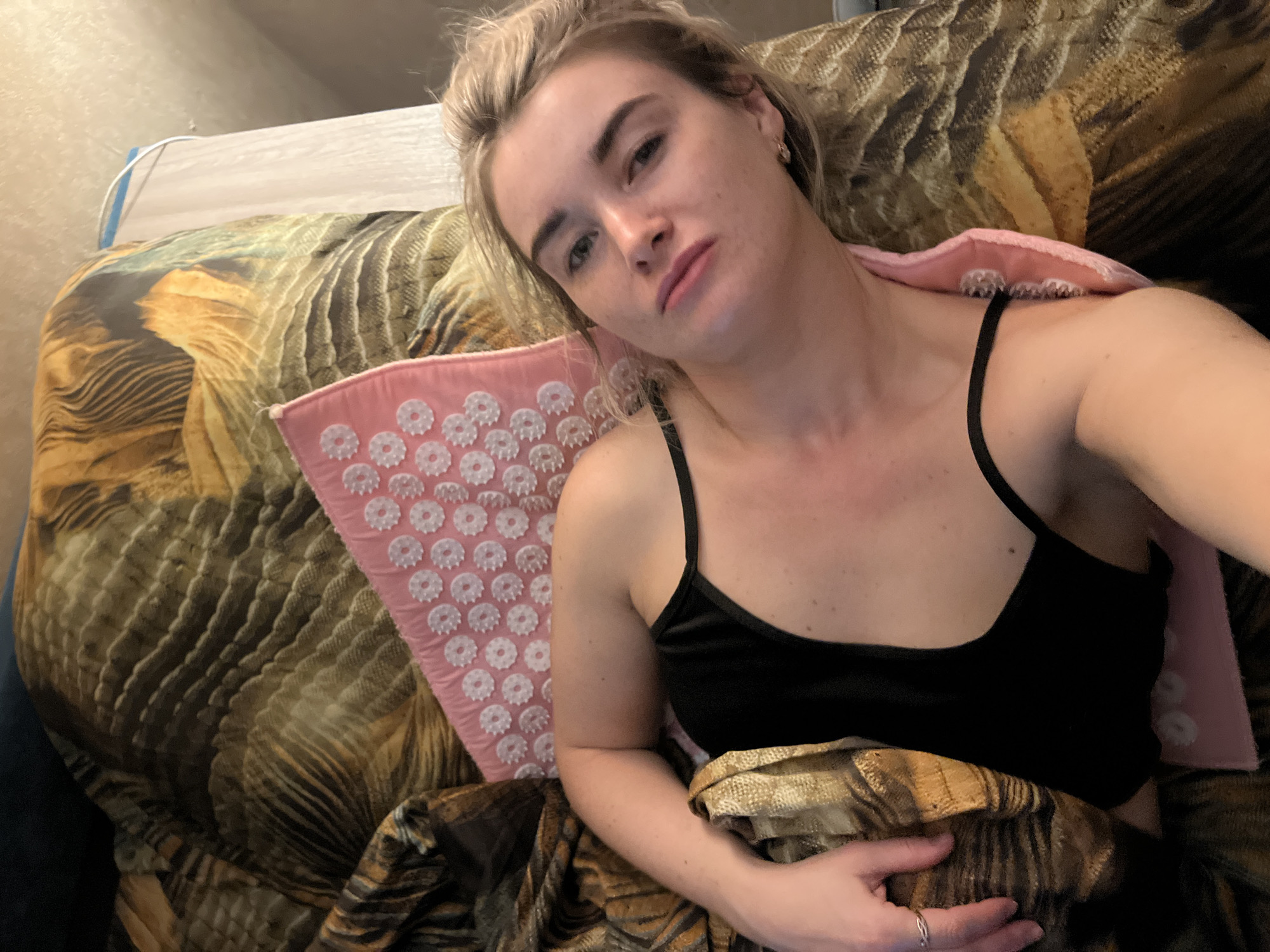 A Caucasian woman with fair skin and blonde hair, possibly in her late 20s to mid-30s, lies reclined, presumably in a residential setting. She wears a black tank top and a ring on her left hand, visible as she rests her hand on a patterned blanket. She appears to be lying on a brown, patterned couch or bed with a pink acupressure mat placed behind her. The overall lighting is soft, suggesting an indoor environment. The location in the city of Chaikovsky, Russia is assumed based on contextual information not available in the image itself.
