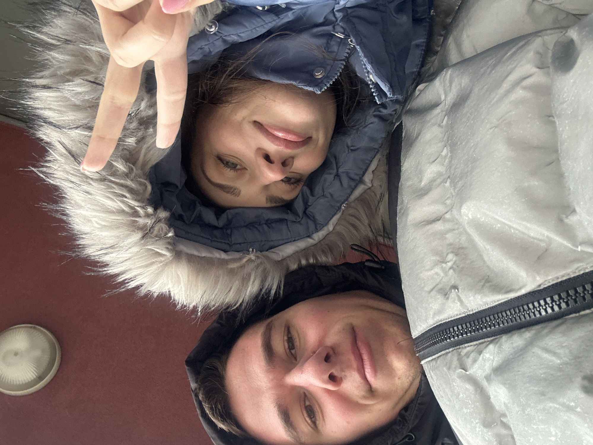 Two people are taking a selfie outdoors. The person on the left is wearing a blue hooded jacket with fur trim and is holding up two fingers. The person on the right is wearing a black hooded jacket and is smiling. A light fixture is visible in the background. It is difficult to determine from this image if the location is Sofia, Bulgaria. More context would be needed.