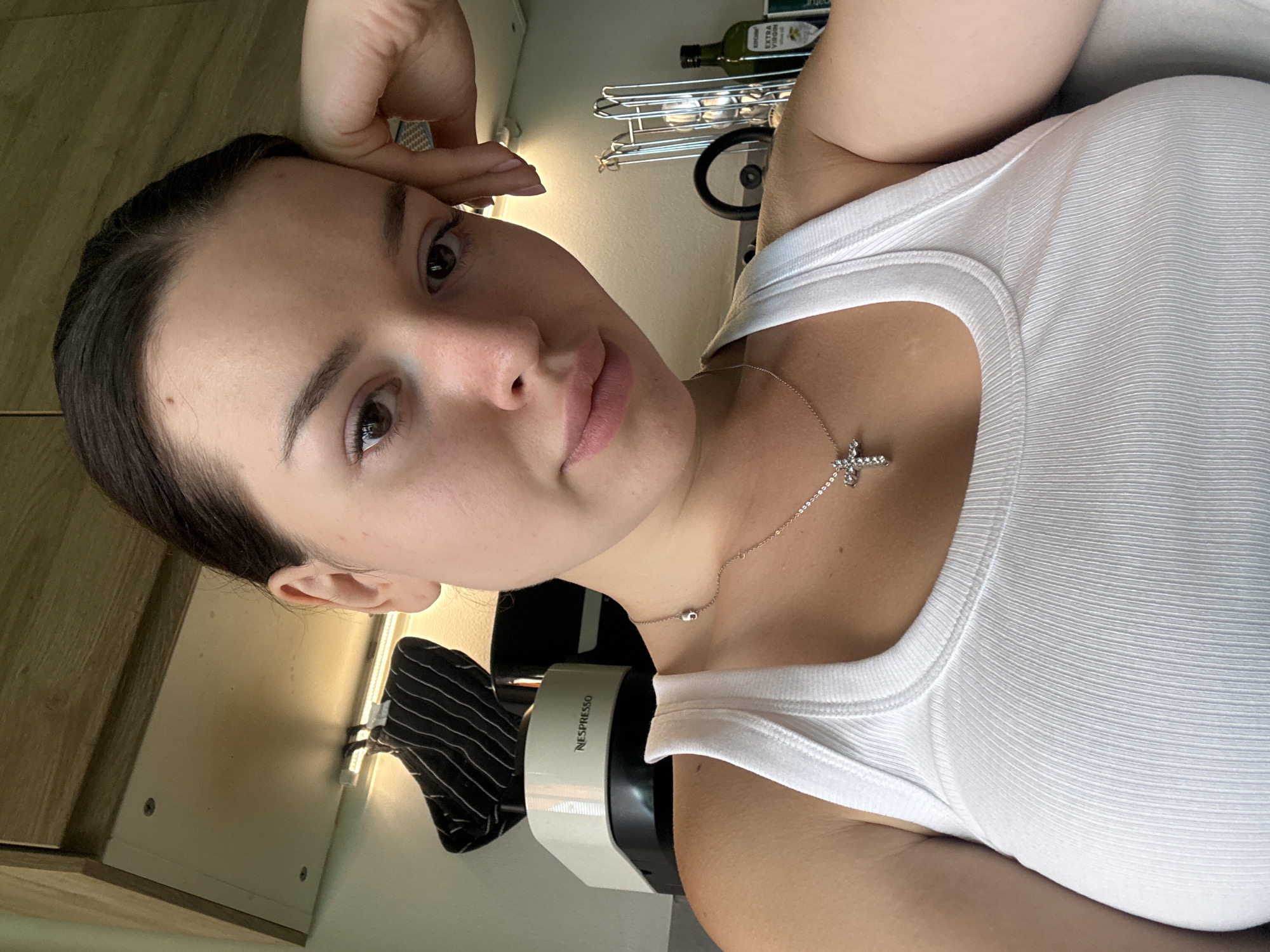 A young woman with dark hair and brown eyes is captured in a close-up selfie, resting her head on her right hand. She is wearing a white ribbed tank top and a silver necklace with a cross pendant. The background suggests a domestic setting, possibly a kitchen, with a Nespresso machine and cabinets visible. The lighting appears to be natural, casting soft shadows on her face. The overall impression is casual and relaxed, with the woman's gaze directed towards the camera.