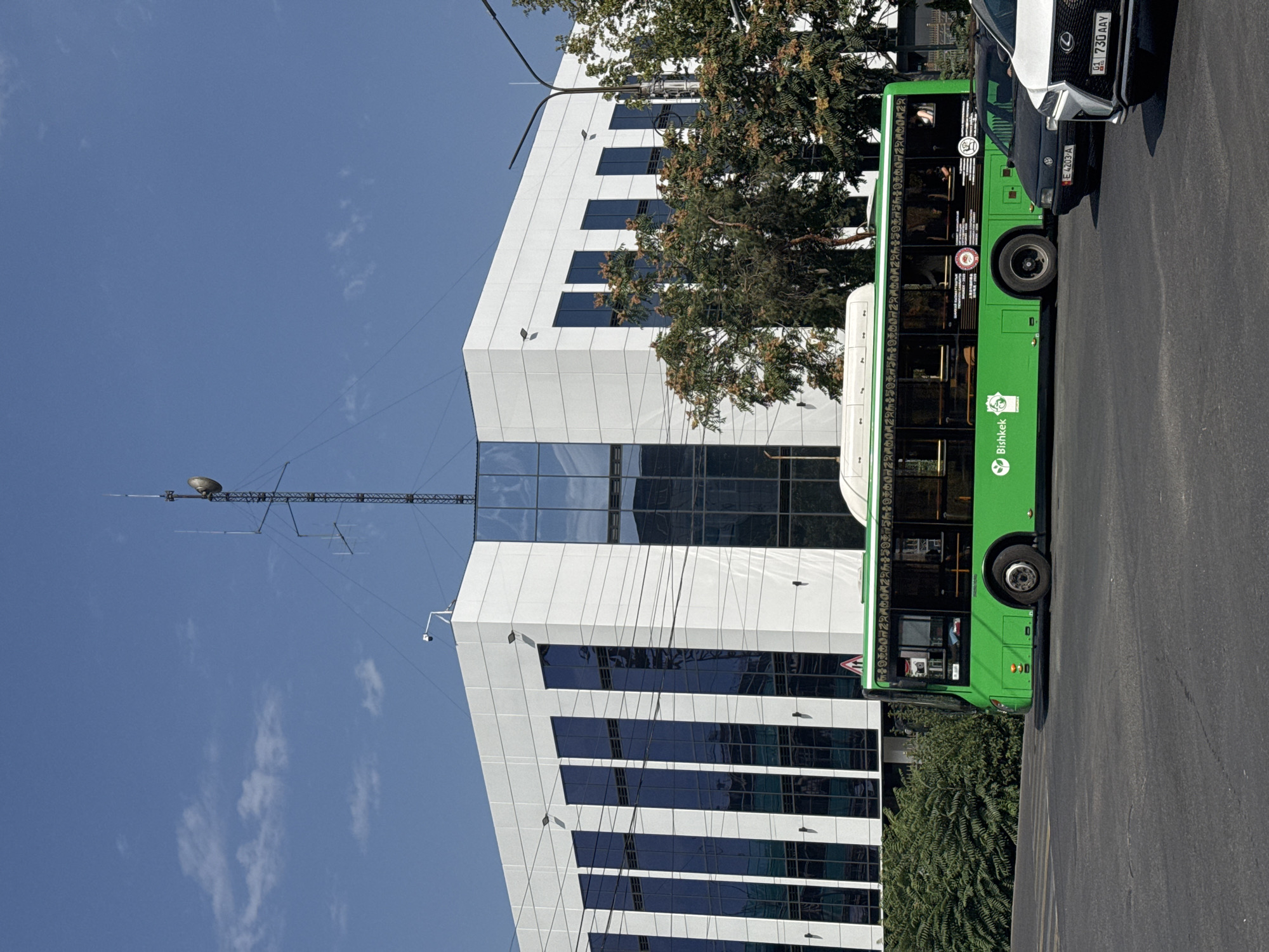 The image shows a modern, white, multi-story building with dark tinted windows and a prominent antenna tower on its roof. A bright green bus with "Bishkek" written on its side is parked in front of the building, on a city street. Several cars are also visible on the road. The scene appears to be in Bishkek, Kyrgyzstan, given the text on the bus. The sky is clear blue with a few wispy clouds, and trees with autumn foliage are visible to the right of the building.