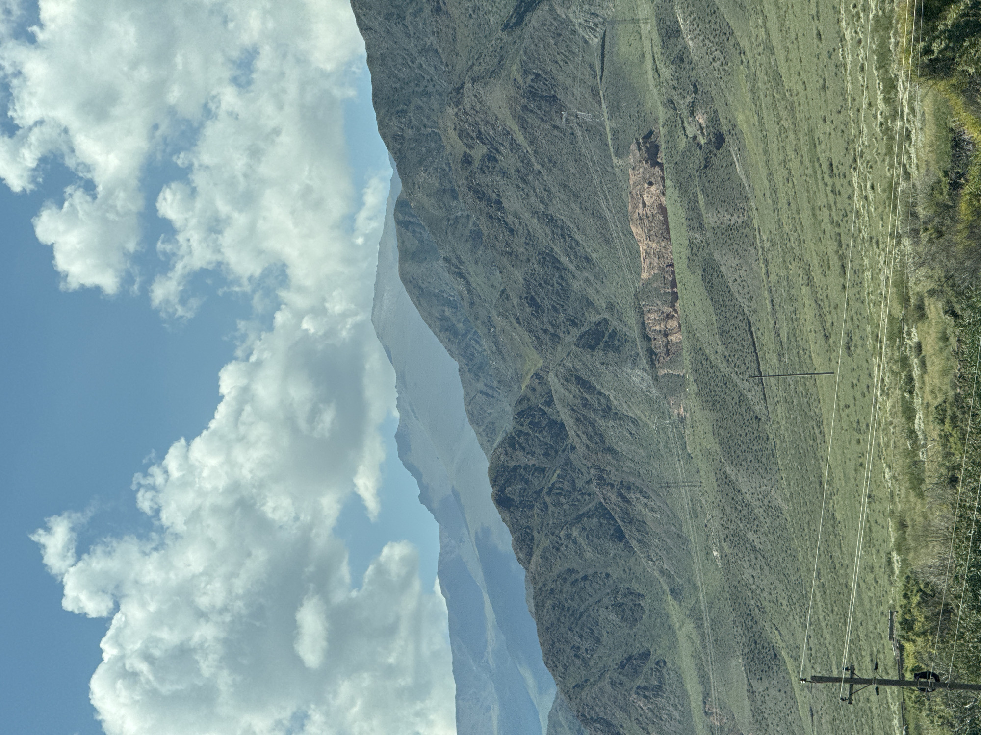 A wide shot captures a mountainous landscape under a partly cloudy sky. The foreground features a grassy plain with scattered bushes and a wooden utility pole supporting power lines. In the mid-ground, the slopes of the mountains are covered in green vegetation, with a visible scar of exposed red rock on one mountainside. The background consists of several layers of mountains, receding in shades of blue and gray, with hazy peaks disappearing into the distance. The sky is a vibrant blue, dotted with fluffy white clouds. No people or specific actions are depicted. The scene appears to be a natural, undeveloped environment.