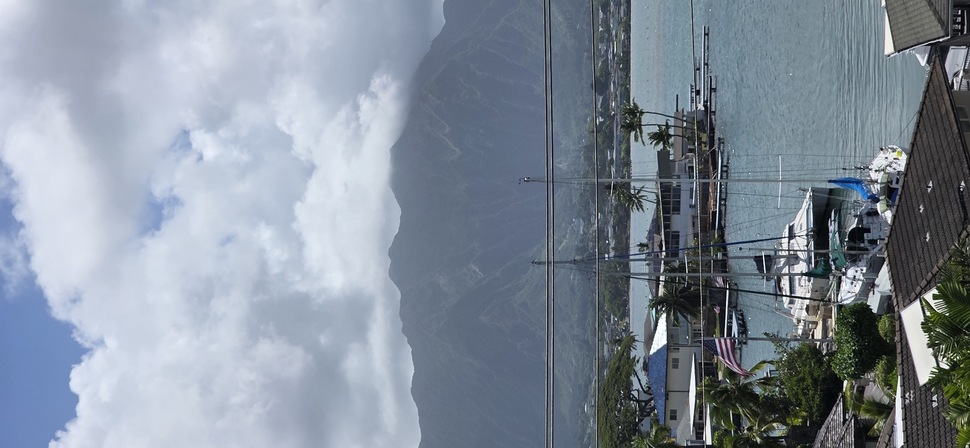 The image depicts a scenic view of Kaneohe, Hawaii, United States of America. In the foreground, several boats are docked in the clear blue water. Among them, a white catamaran stands out with its distinctive hull and multiple masts. Behind the boats, residential buildings with tiled roofs and palm trees line the shore. An American flag is prominently displayed on a flagpole near one of the houses. In the mid-ground, more buildings and vegetation are visible, suggesting a developed coastal area. The background is dominated by a majestic mountain range, partially shrouded in clouds, characteristic of Hawaii's lush volcanic landscape. The sky above is a mix of blue patches and fluffy white clouds, indicating a partly cloudy day. Several power lines stretch across the upper portion of the image, a common urban element. The overall scene conveys a tranquil, tropical atmosphere with a blend of natural beauty and human settlement.