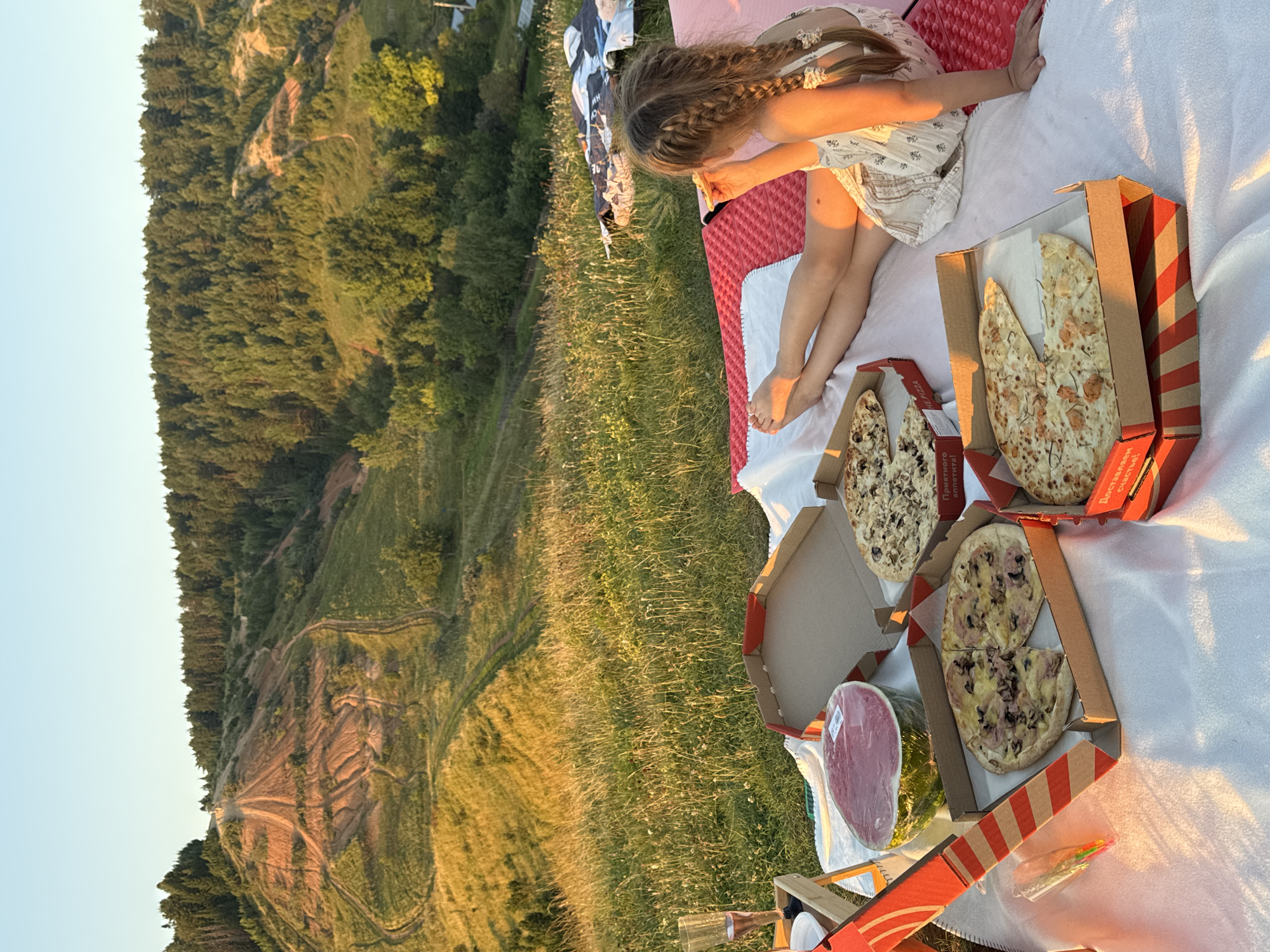 A young girl with long, braided hair is having a picnic outdoors. She is wearing a light-colored dress with a floral pattern. She is sitting on a white blanket spread over a red mat on a grassy hillside. Beside her are several open pizza boxes containing various pizzas. A large watermelon wrapped in plastic is also on the blanket. In the background, a wooded hillside with a winding path is visible, bathed in the warm light of the setting sun. The sky is a clear, pale blue. The overall scene suggests a peaceful, late afternoon or early evening picnic in a rural or natural setting, possibly in Olkhovka, Russia. Some text is visible on the pizza boxes, including "ПРИЯТНОГО" and "СПАСИБО".