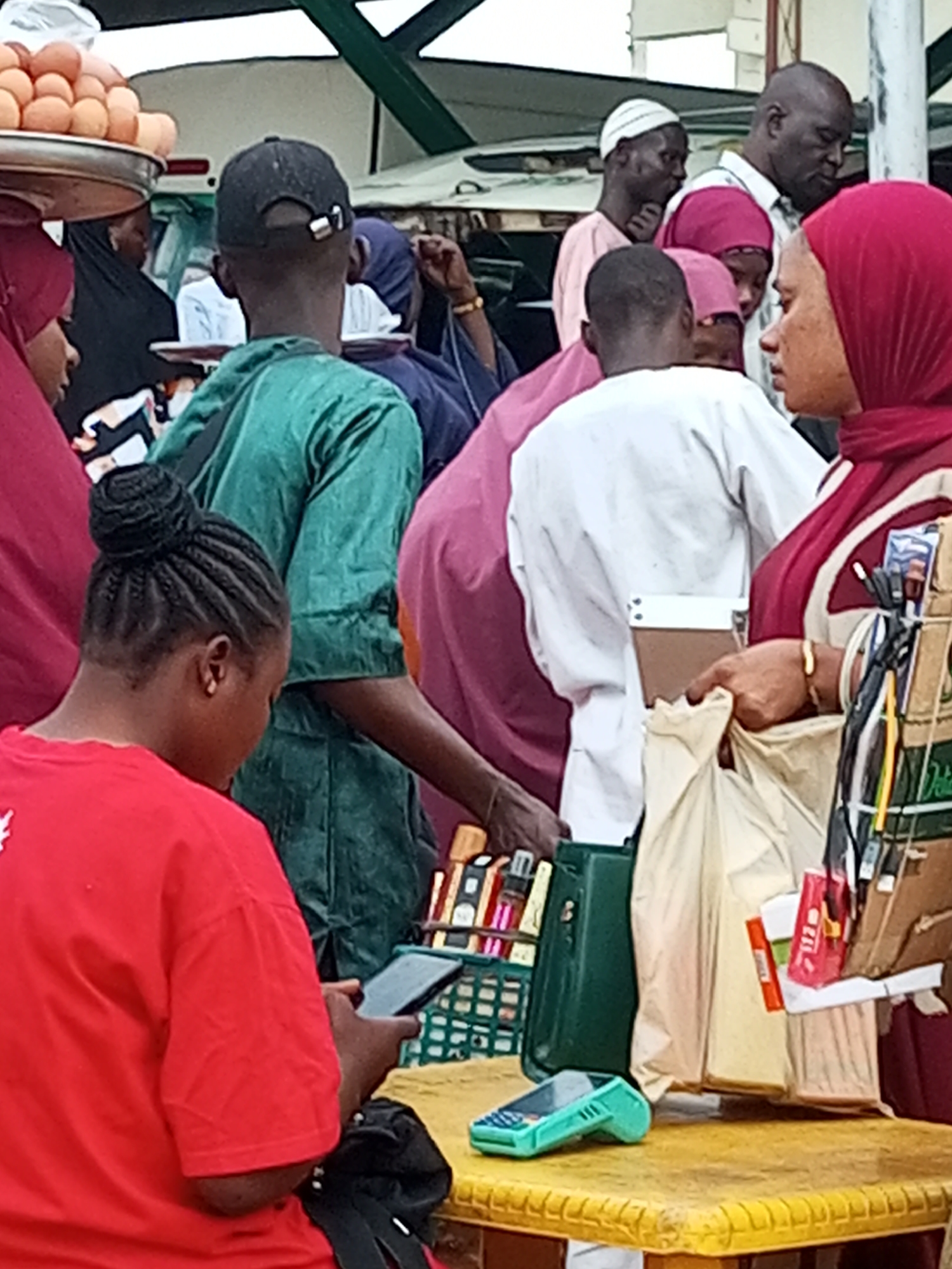 This image, captured in the city of Gombe, Nigeria, depicts a bustling marketplace scene with several individuals engaged in various activities. In the foreground, a person in a red t-shirt with their hair in braids is looking down at a smartphone, which they are holding in their left hand. To their right, a woman wearing a red hijab and a maroon and cream patterned garment is interacting with a collection of items displayed on a table, including bags of merchandise and what appears to be a credit card reader. Behind them, a man in a green t-shirt and a black baseball cap is visible, along with other individuals wearing hijabs in various colors. In the background, a stall with a large quantity of eggs on a metal tray is partially visible, suggesting the sale of food items. The overall environment appears to be an outdoor market with a covered structure in the background, likely a vendor's stall. The lighting suggests it is daytime.