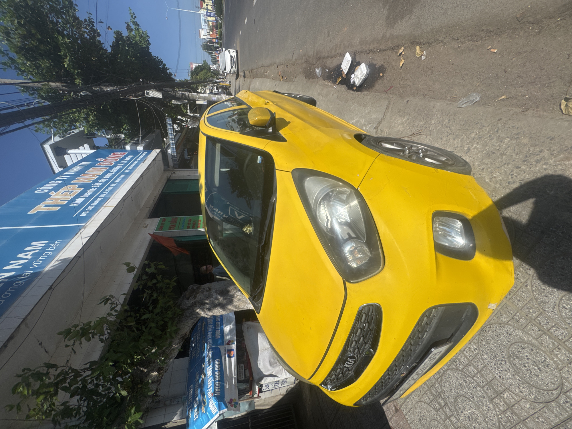 A bright yellow Kia car is parked on the side of a street in the city of Bac Nha Trang Ward, Vietnam. The car is facing towards the right side of the image, with its front quarter visible. The car's hood, headlights, and a portion of its side are in clear view. It is parked on a paved sidewalk next to a street where other vehicles are visible in the distance.
To the left of the car, there is a building with blue and white signage, some of which has text in Vietnamese. A tree with green leaves is growing next to the building, partially obscuring the signage. Behind the car, a person wearing a light-colored shirt is visible, standing near the building.
The street appears to be a typical urban road with power lines and utility poles running along its side. There is some debris on the ground near the car, including plastic bags and leaves, suggesting a public area. The sky is clear and blue, indicating a sunny day. The overall scene captures a moment of everyday life in a Vietnamese urban environment.