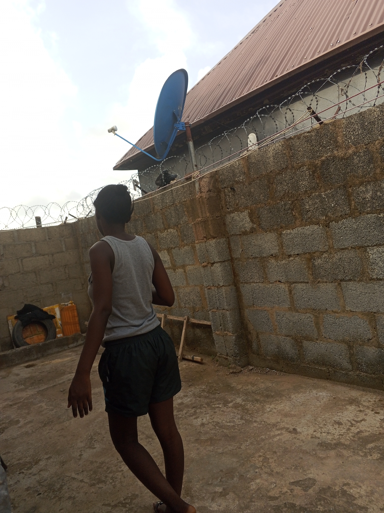 A young person with dark skin wearing a gray tank top and dark shorts stands in a walled, sunlit courtyard in Abuja, Nigeria. They are facing away from the camera. The courtyard is enclosed by cinder block walls topped with barbed wire. A blue satellite dish is mounted on a building with a brown corrugated metal roof visible behind the walls. In the corner of the courtyard, there is a tire and some covered objects on a concrete platform. A wooden ladder leans against the wall. The ground is bare concrete.