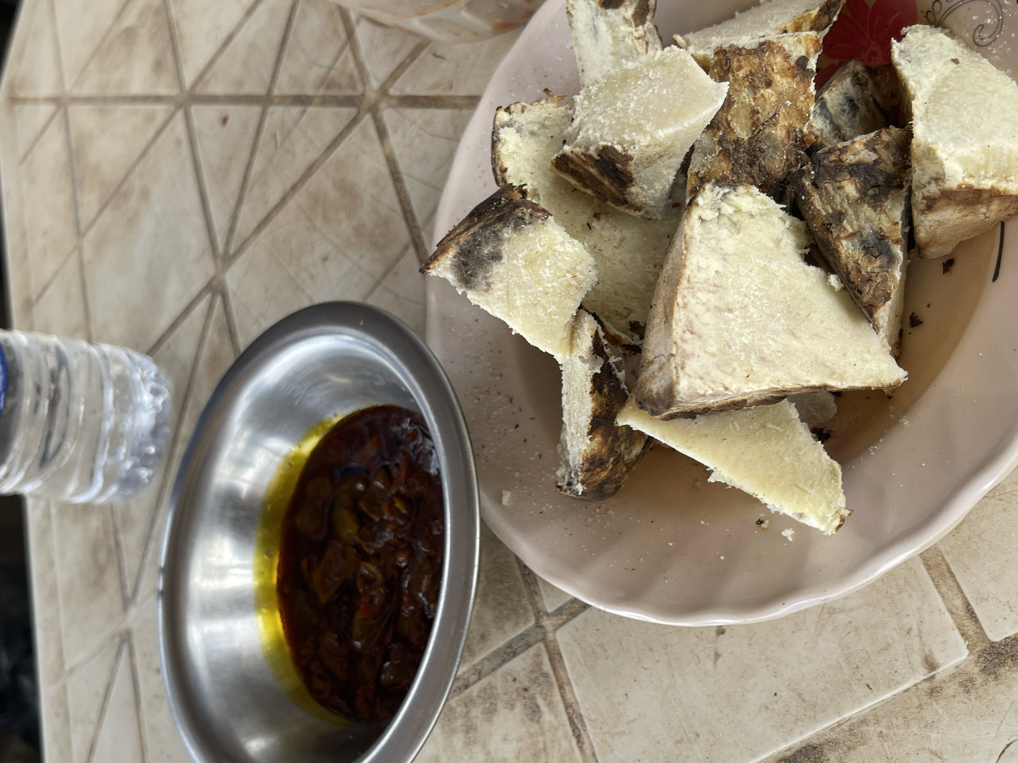 The image shows a plate of roasted yam served with a sauce, likely photographed in Angwan Sayawa, Nigeria. The yam is cut into triangular pieces with dark brown/blackened edges. The sauce is in a small, round, stainless steel bowl and appears to be a dark red/brown color with an oily yellow perimeter. A bottle of water is on the left. The dishes are set on a tiled surface that resembles a table.