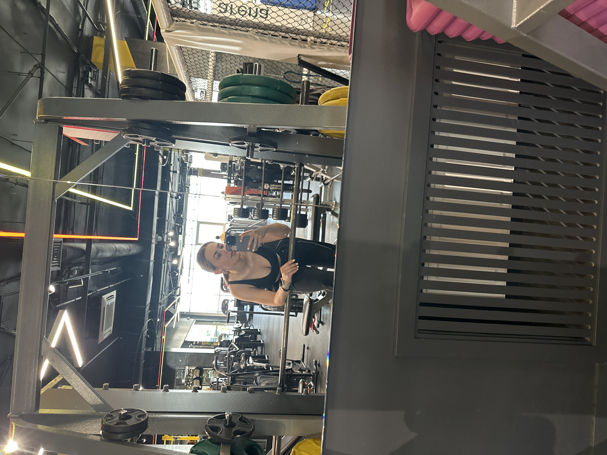 A woman with blonde hair pulled back, wearing a black tank top and black leggings, takes a mirror selfie in a gym. She is holding a smartphone in her right hand. She is leaning on a weight rack with her left arm. The gym appears to be modern and well-equipped, with various weight machines and equipment visible in the reflection. The lighting is bright, with some neon accents on the ceiling. The floor is dark, and there is a large metal vent on the lower part of the image. Based on the provided information, this image was taken in the city of Perm, Russia.
