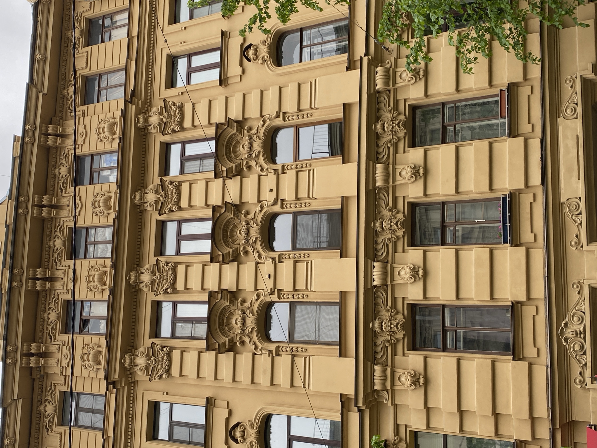 Exterior view of a tall building facade in Saint Petersburg, Russia. The building is tan in color and features elaborate decorative moldings and window frames. The windows are rectangular and arched. Some green foliage is visible at the top of the building. The sky is overcast.
