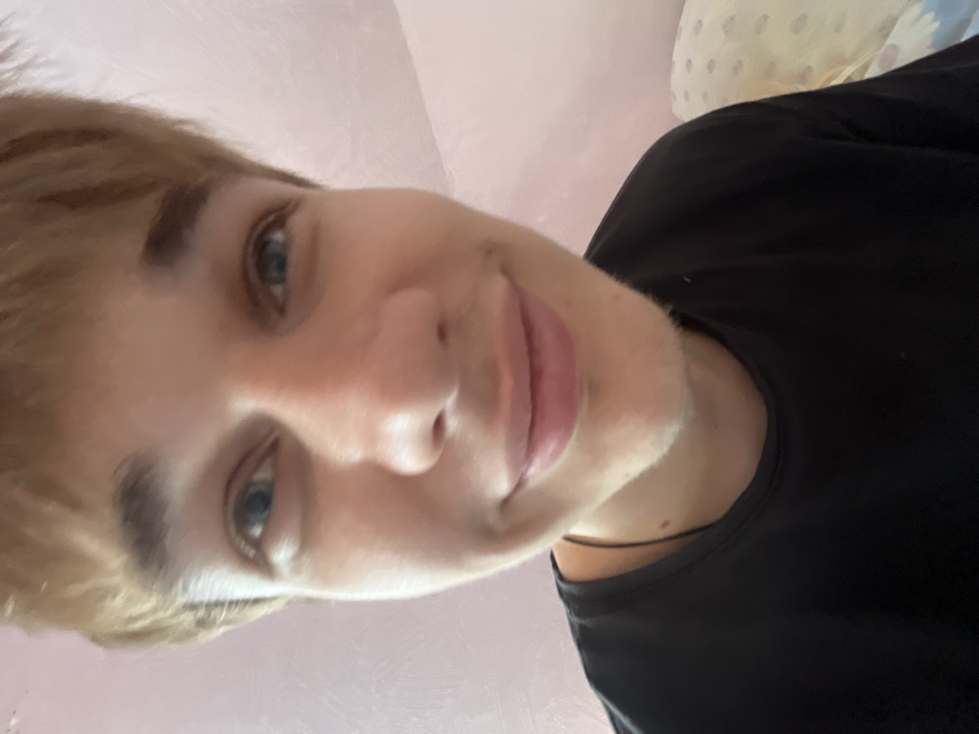 The image is a close-up, fisheye-lens selfie of a young Caucasian male with light brown hair and blue eyes. He is wearing a black t-shirt and appears to be indoors, with a pink wall in the background. There is no indication of the location in the city of Gari, Russia. The subject is looking directly at the camera with a slight smile. The lighting suggests it is daytime.
