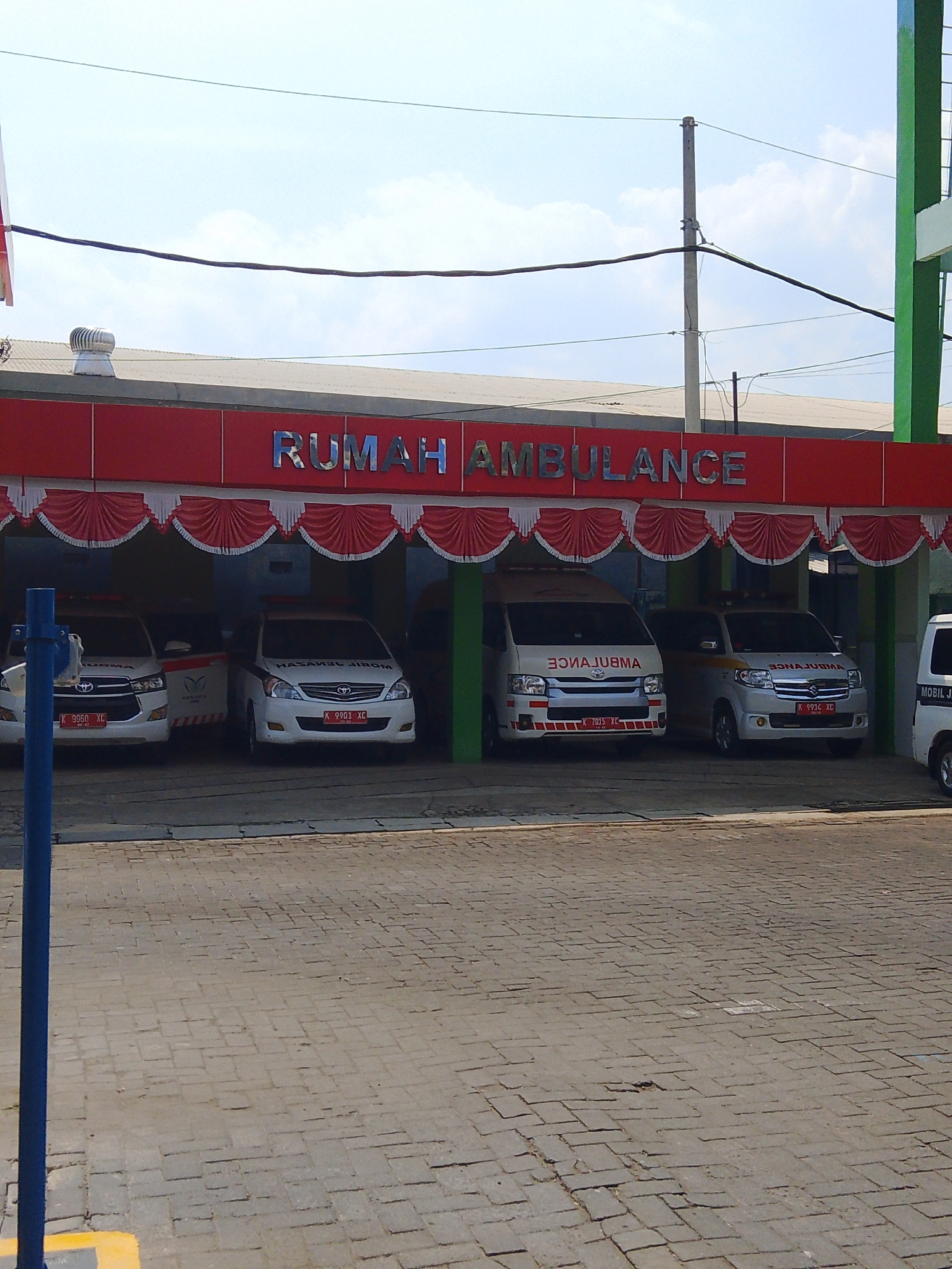 The image depicts an ambulance station, labeled "RUMAH AMBULANCE," situated in Potroyudan, Indonesia. The building features a red facade with a white and red scalloped valance above the entrance. Inside the open bays, four white ambulances are parked. Two of the ambulances are visible on the left, and two on the right. The ambulance on the far right is partially obscured. The ground in front of the station is paved with grey interlocking bricks. A blue pole with a yellow marking stands in the foreground on the left. The sky above is a clear light blue.