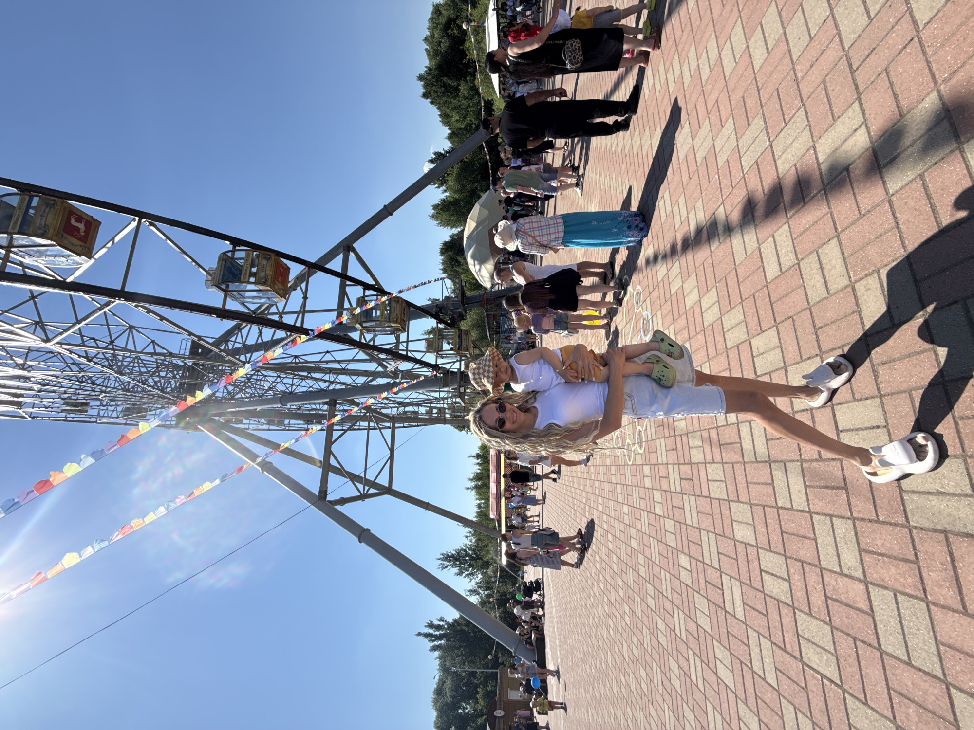 An outdoor, low-angle, wide shot captures a woman holding a toddler at an amusement park, in front of a Ferris wheel. The woman, in her late 20s, has long blonde hair, sunglasses, a white tank top, and denim shorts. She is holding a toddler, wearing a checkered hat and shorts, in her arms. The background is a paved area with people milling around, a Ferris wheel, and trees. The sky is clear and blue, with the sun casting long shadows. The scene is bright and sunny.