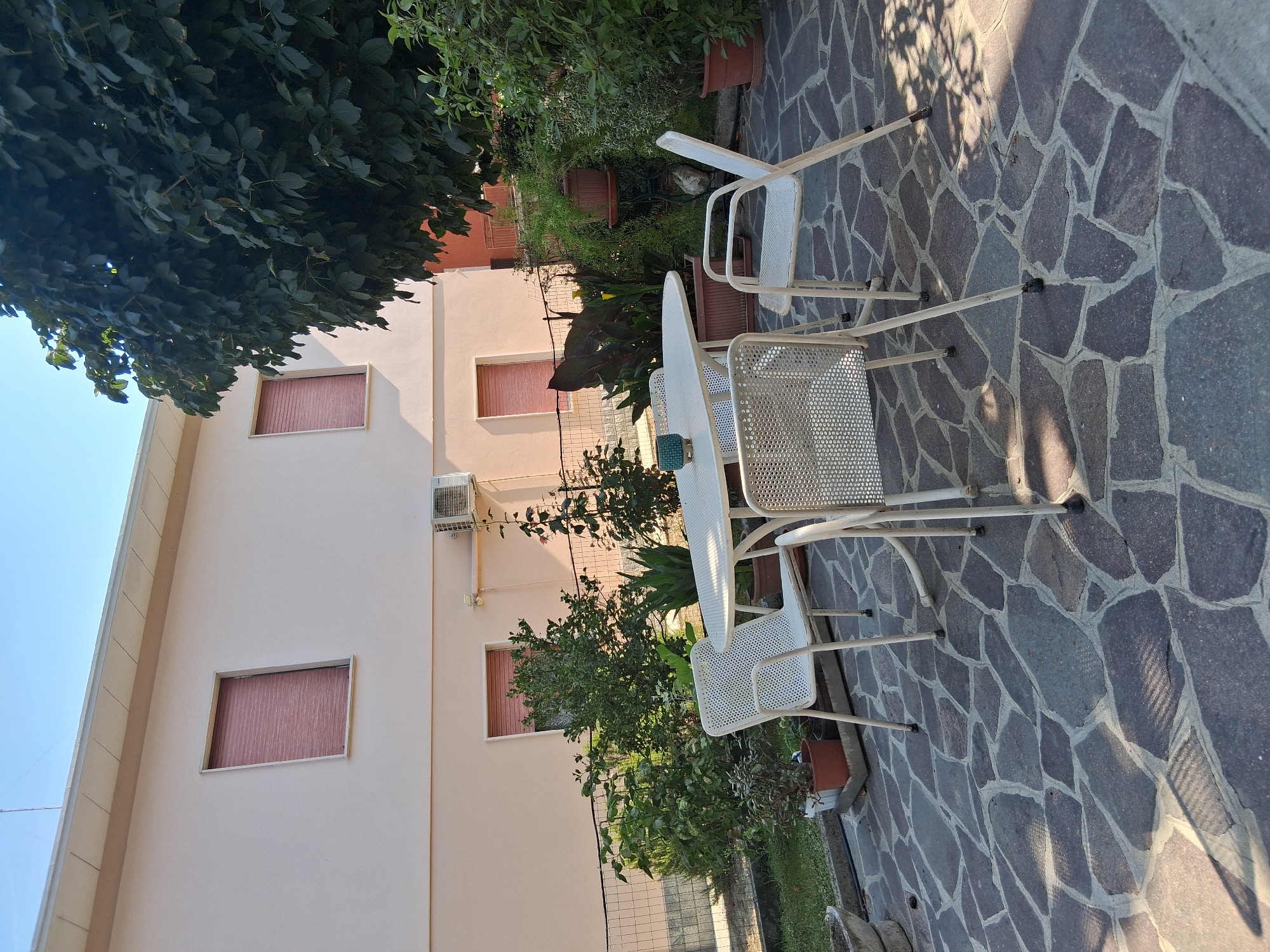 The image depicts a sunny day in the city of Colorno, Italy, in a residential backyard. A pale pink building with two windows that have red roller shutters is visible in the background. To the right of the building, a large tree with dark green leaves casts shadows on the scene. In the foreground, a patio is paved with irregularly shaped gray stones. A white oval table with four white perforated metal chairs is set up on the patio. The table has a small blue decorative object on it. Around the patio, there are various potted plants and a small garden area separated by a metal fence. The overall atmosphere is peaceful and suggests an outdoor seating area for relaxation or dining.