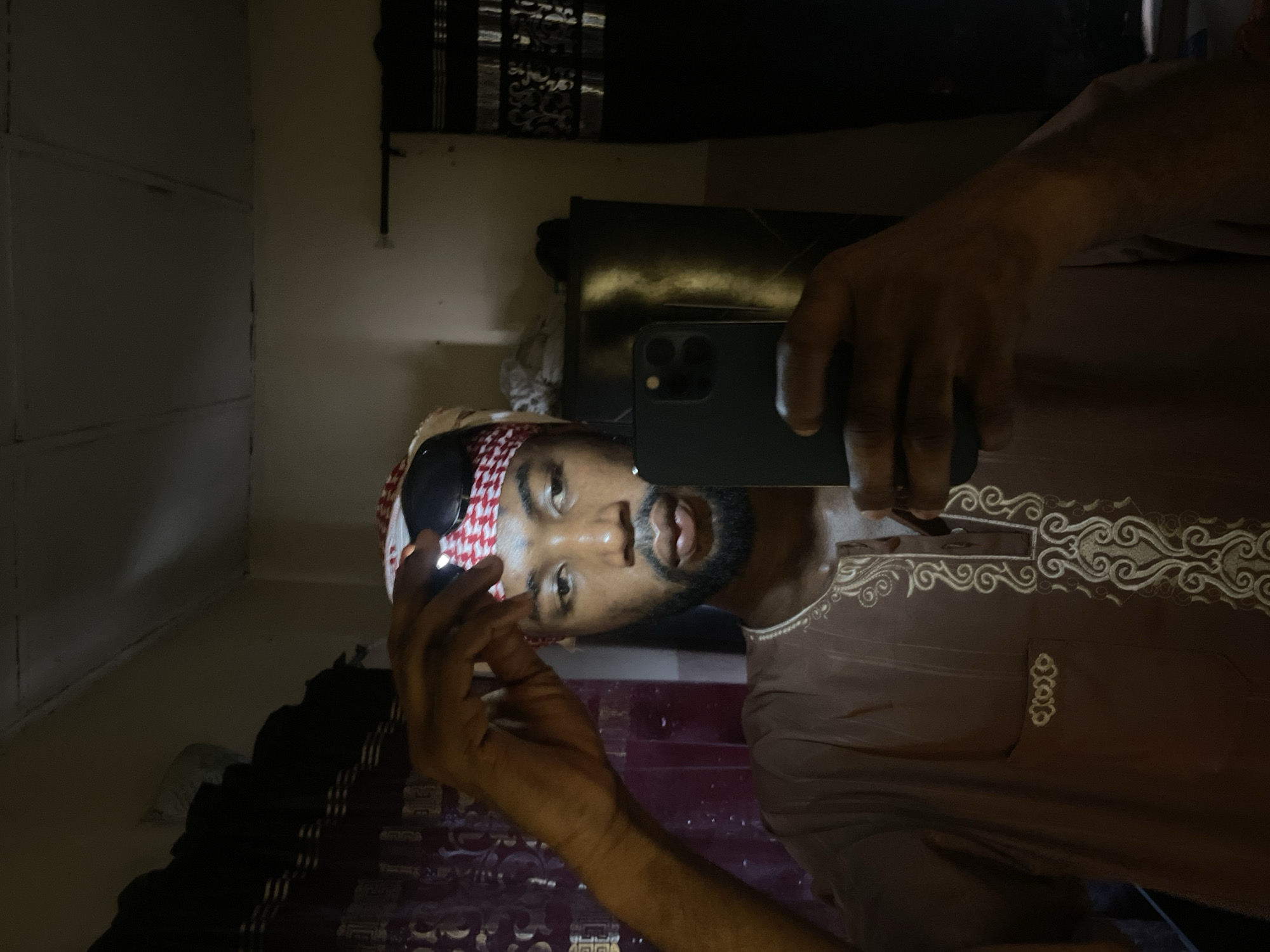 A man wearing a red and white patterned headscarf and sunglasses is taking a selfie with a dark-colored smartphone. He is wearing a brown shirt with decorative embroidery around the collar and down the front. The background is a dimly lit room with a partially visible dark-colored cabinet or dresser. The location in Lokoja, Nigeria is not determinable from the image.