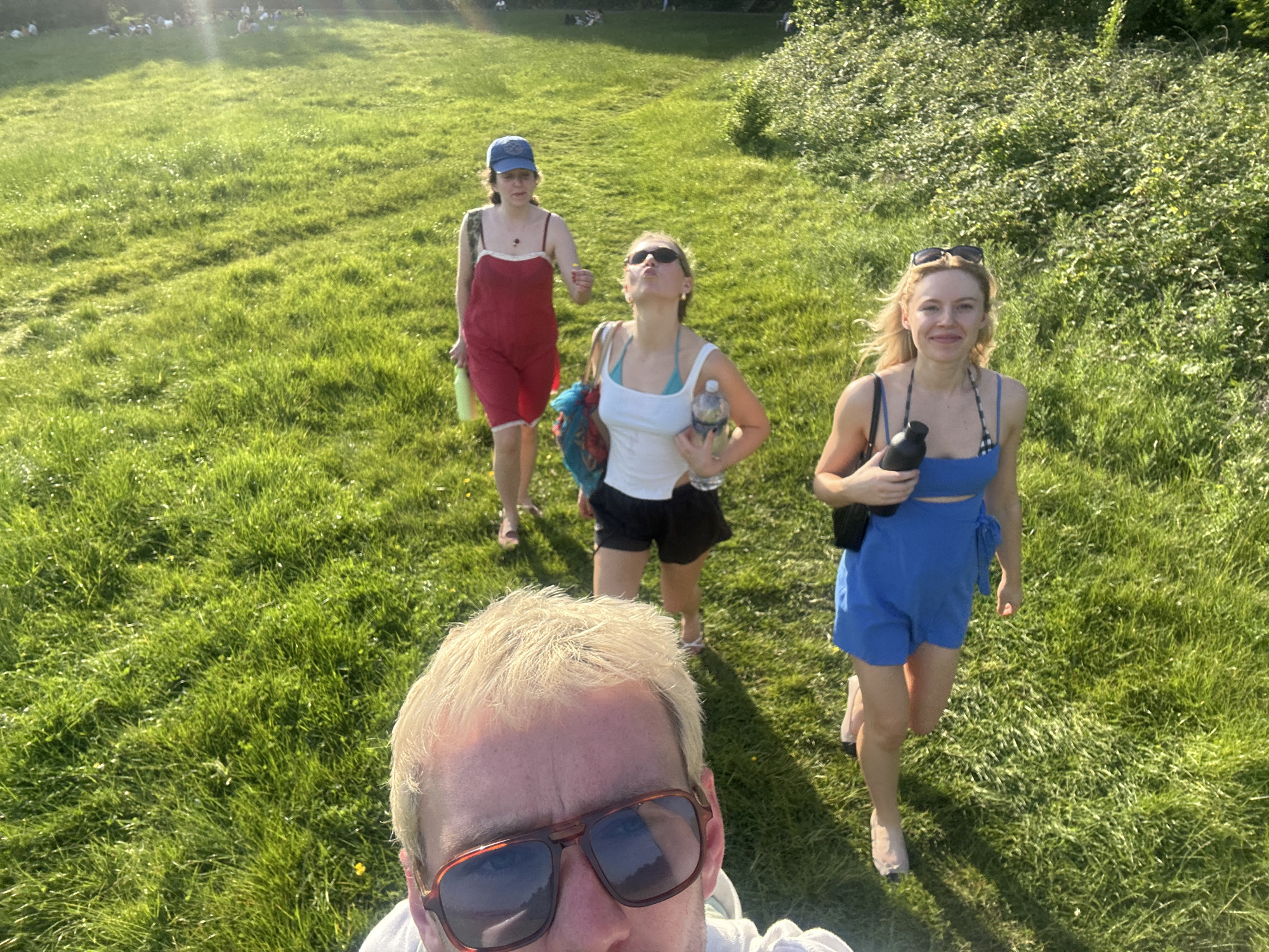 The image is a selfie taken outdoors, likely in a park in London, United Kingdom, given the lush green grass and the presence of people in the background, suggesting a recreational area. The foreground is dominated by a person with short, blond hair and sunglasses. Behind the person taking the selfie are three women walking towards the camera. One woman is wearing a red romper with a blue baseball cap and carrying a green bottle. Another is wearing a white tank top and black shorts, with sunglasses and a water bottle. The third woman is wearing a blue romper and holding a black water bottle. The lighting suggests it is daytime, with a bright sun. The background shows a grassy field with people sitting and relaxing. The environment is green, open, and natural.