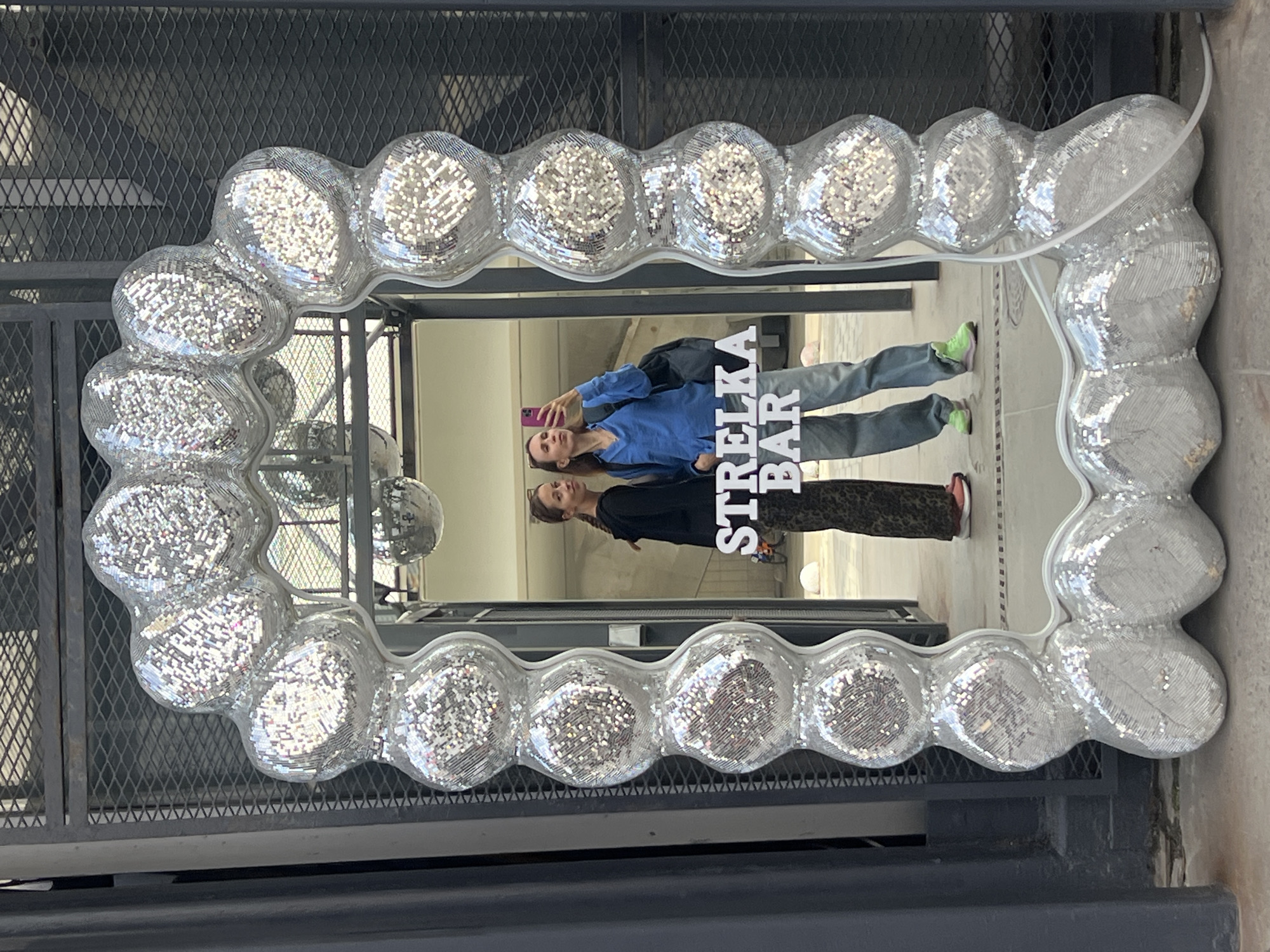 A rectangular mirror with a frame composed of interconnected, disco ball-like spheres is displayed against a wrought-iron fence. The frame's material gives a mirrored, fragmented reflection. The mirror's reflection shows two women taking a selfie with a smartphone, and the words "STRELKA BAR" in white capital letters. The background visible in the reflection suggests a public, urban environment. Given the text, the image is likely located in Moscow, Russia. The mirror is outside, suggesting it might be part of a public art installation or advertising for the Strelka Bar.