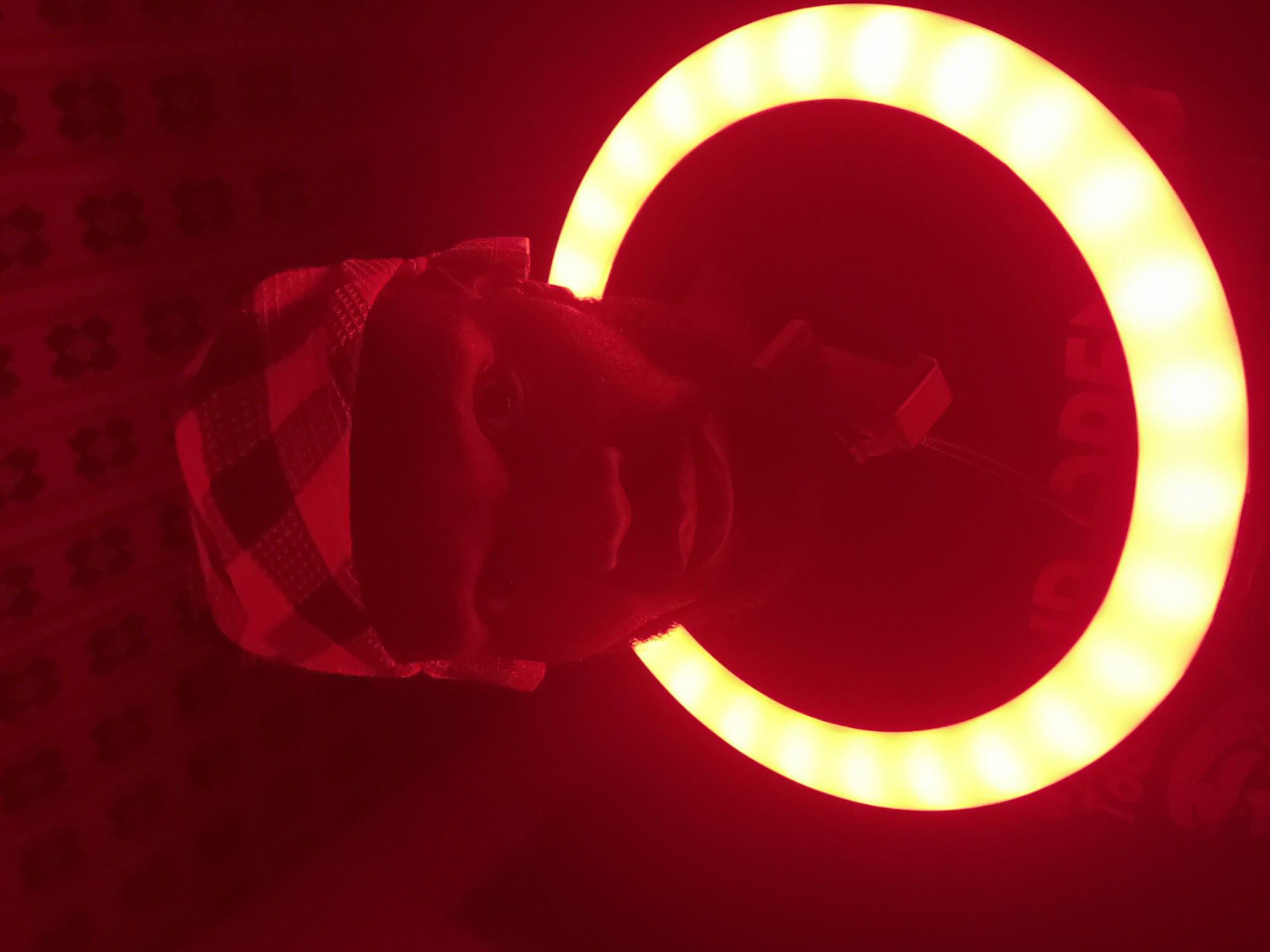 The image depicts a person illuminated by a vibrant, circular LED ring light against a dark red background. The scene is set in Gura Top, Nigeria. The subject, likely a young adult, is positioned centrally, with their face partially obscured by the intense yellow-white glow of the ring light. They are wearing a patterned head covering, possibly a checkered scarf, that is red and darker in tone.
The most prominent object is the large ring light, emitting a bright, warm yellow-white illumination that outlines the subject's head and shoulders. A small black bracket, holding what appears to be a smartphone or compact camera, is attached to the lower right side of the ring light, indicating a setup for taking photos, selfies, or recording video. Faint text spelling "MODE" is visible on the black casing of the ring light near its bottom right curve.
The background is a deep, uniform red, suggesting an indoor setting with red ambient lighting or a red wall/fabric. A subtle, darker red floral or geometric pattern is faintly discernible on the background, adding texture. The overall lighting scheme, combining deep red with the bright ring light, creates a dramatic and focused visual. The activity appears to be content creation or a video call, utilizing the specialized lighting equipment in a low-light environment, likely at night. No specific emotions are clearly visible due to the heavy backlighting and limited view of the face.