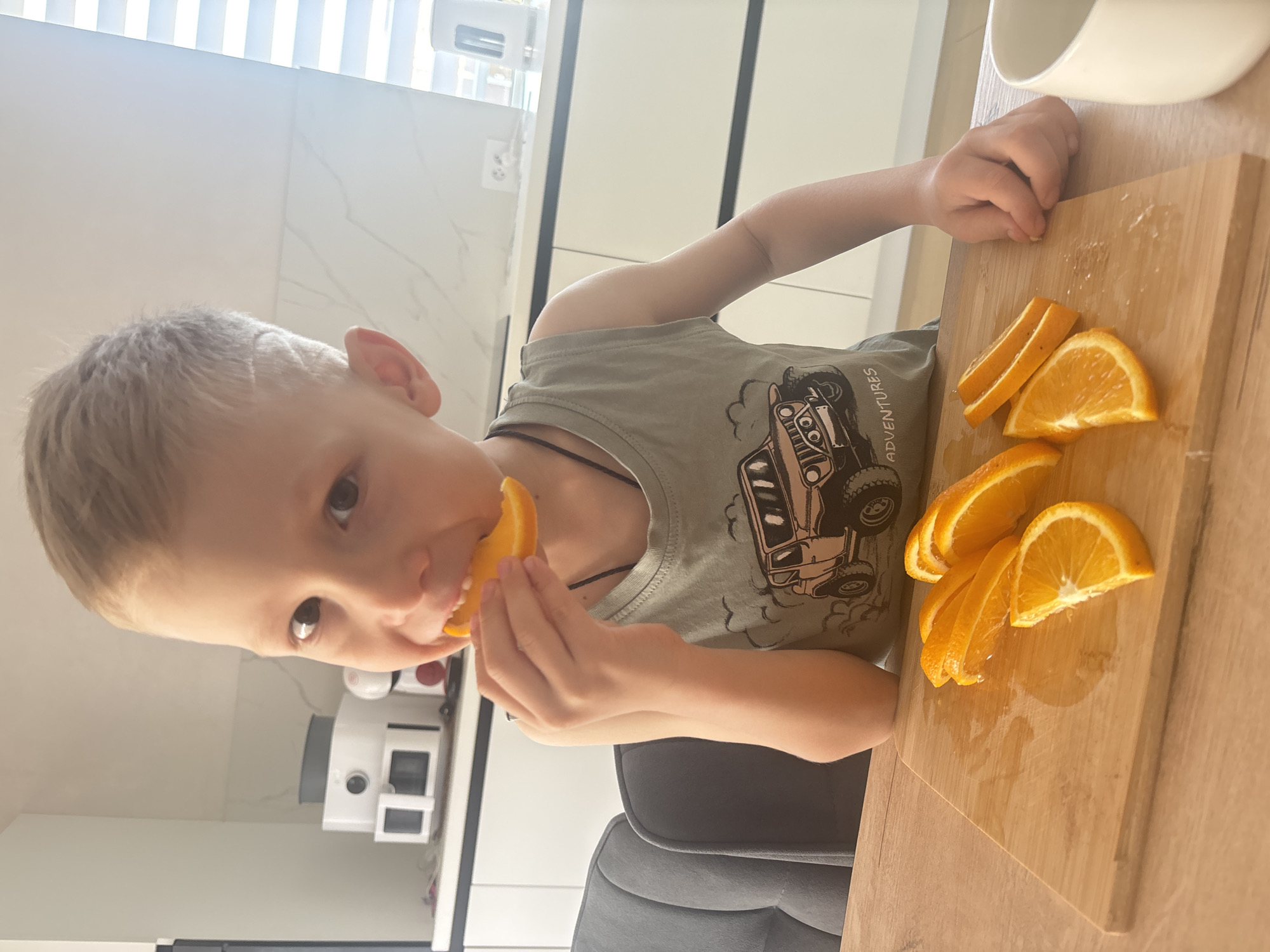 A young boy is seated at a table, eating a slice of orange. He is wearing a light green tank top with a vehicle graphic. A wooden cutting board sits on the table, holding several slices of orange. A white bowl is partially visible at the top of the frame. The scene appears to be indoors, possibly in a kitchen, based on the countertops and appliances visible in the background. The location is unknown, but Russia is included as the origin.