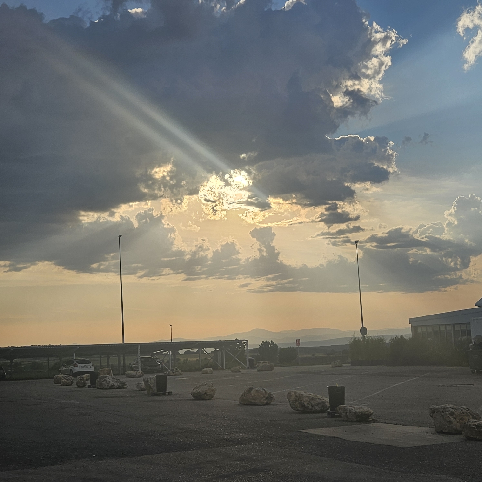 The image depicts a parking lot scene in Algora, Spain. The foreground shows a paved parking area with several large, light-colored rocks scattered throughout. Two metal trash receptacles are also present. In the background, a covered parking structure is visible, along with distant rolling hills or mountains. Two light poles with streetlights are seen. The sky is dominated by thick clouds, with rays of sunlight breaking through. The lighting suggests either sunrise or sunset. There are no people visible in the image. The environment indicates a rural or semi-rural location.