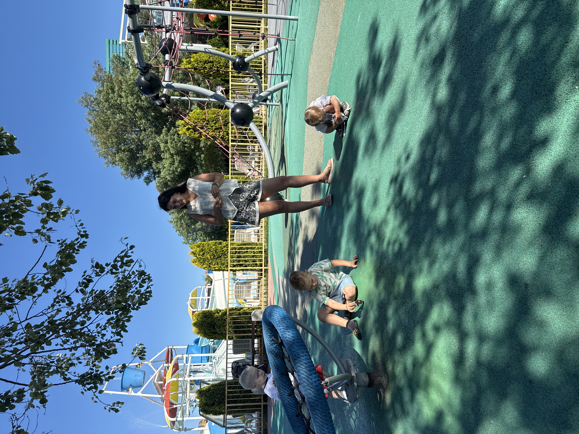 This image depicts a sunny day at a children's playground, likely within the city of Sochi, Russia, given the presence of a water park in the background. A woman stands in the middle ground, looking down at something in her hands. Three young children are playing on the green, rubberized surface of the playground. One child, a boy wearing a green and white striped t-shirt and blue shorts, sits on the ground to the right, playing with a small toy car. Another boy, wearing a white t-shirt and red shorts, stands near a large blue rope structure on the left. A third, smaller child, dressed in a white shirt and striped shorts, is crouched on the ground in the right foreground, also engaged with a toy. The background features elements of a water park, including brightly colored slides and a large blue bucket, suggesting a resort or amusement area. Trees and foliage are visible, along with fencing and part of a climbing structure. The ground is a mottled green, with dappled shadows from overhead trees creating a textured effect.