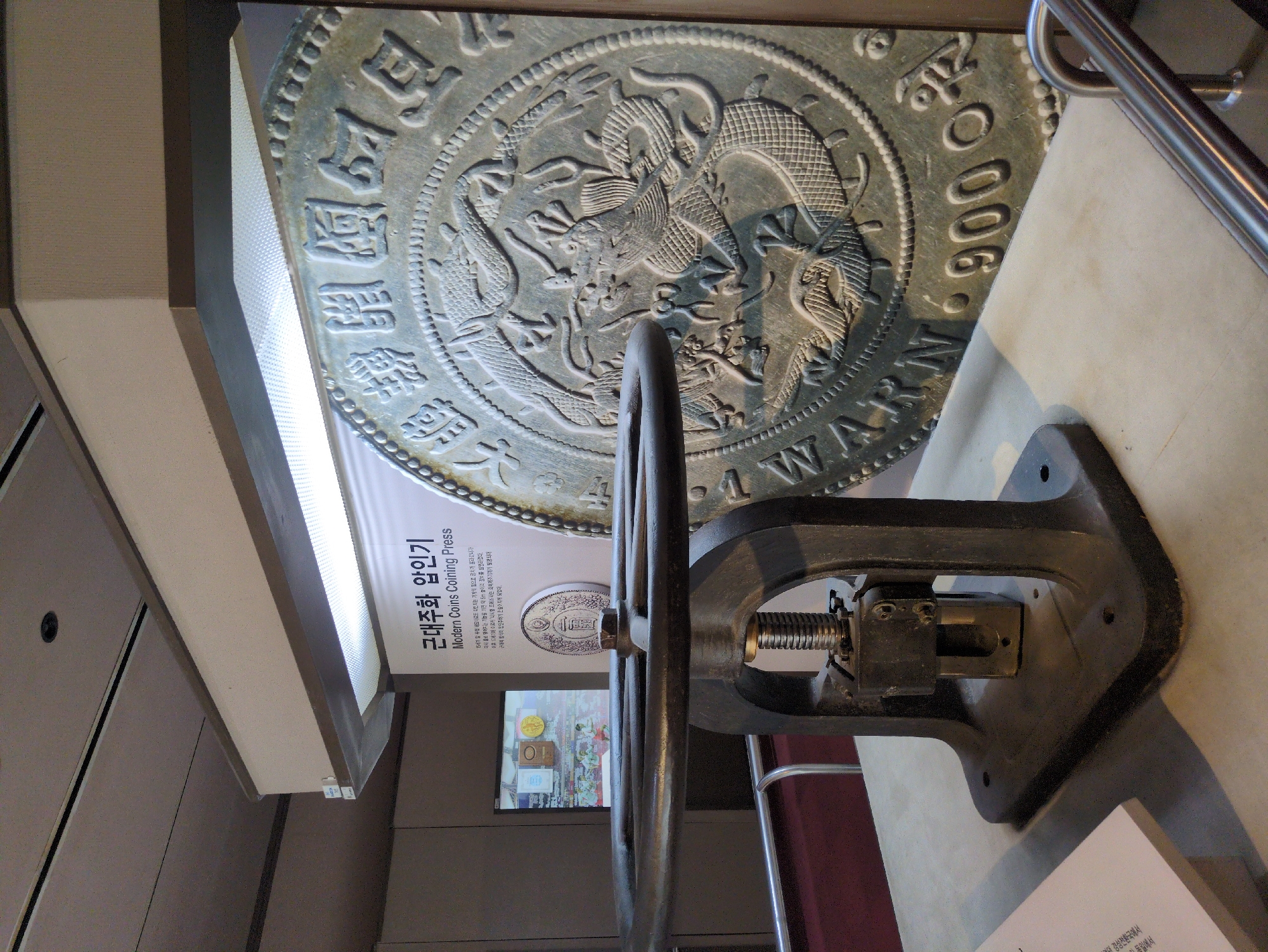 This photograph captures a display of a "Modern Coins Coining Press" at an exhibit in Daejeon, South Korea. A large, metallic, circular relief of a historical coin with dragons and inscriptions dominates the background. The press itself, a heavy iron machine with a large wheel and screw mechanism, occupies the foreground. Text is visible on a sign next to the press, providing information in Korean and English about the coining press. A television screen displaying additional images is visible in the background. The scene suggests a museum or educational setting dedicated to showcasing historical artifacts and technology.