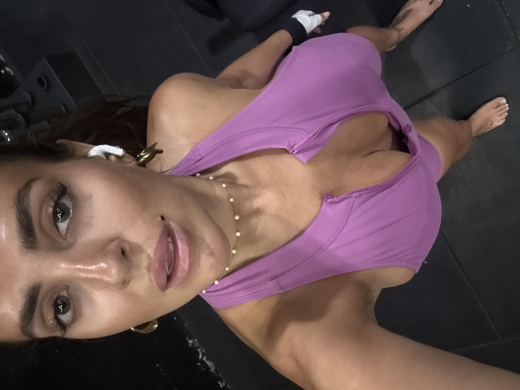 A woman is taking a selfie in what appears to be a gym setting. She is wearing a purple zippered athletic top and a pearl necklace. An earbud is visible in her left ear. Her right wrist is wrapped with white tape over a black band. She is barefoot and standing on a dark, tiled floor. Weights and gym equipment are partially visible in the background. The location is possibly in North Kuta, Indonesia.