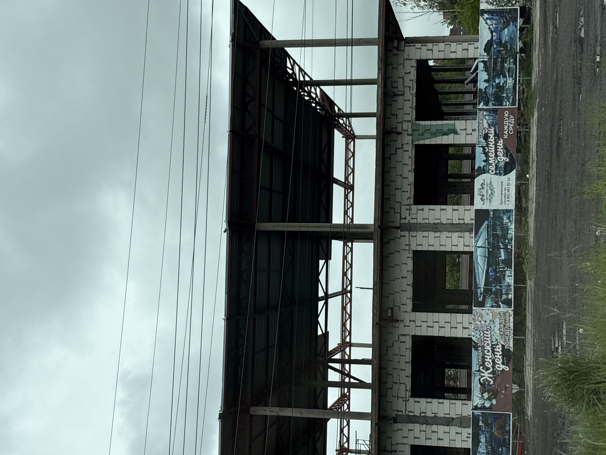 The image shows an unfinished building under construction in what appears to be Novosaratovka, Russia, based on the Cyrillic lettering on nearby advertising banners. The building has a metal roof structure, concrete supports, and walls constructed from white blocks with window openings. Power lines are visible in the sky. Adjacent to the building is a series of banners advertising "Women's Day" and "Family Day" discounts, indicating local business promotions. A road and some grass are in the foreground to the right of the building.
