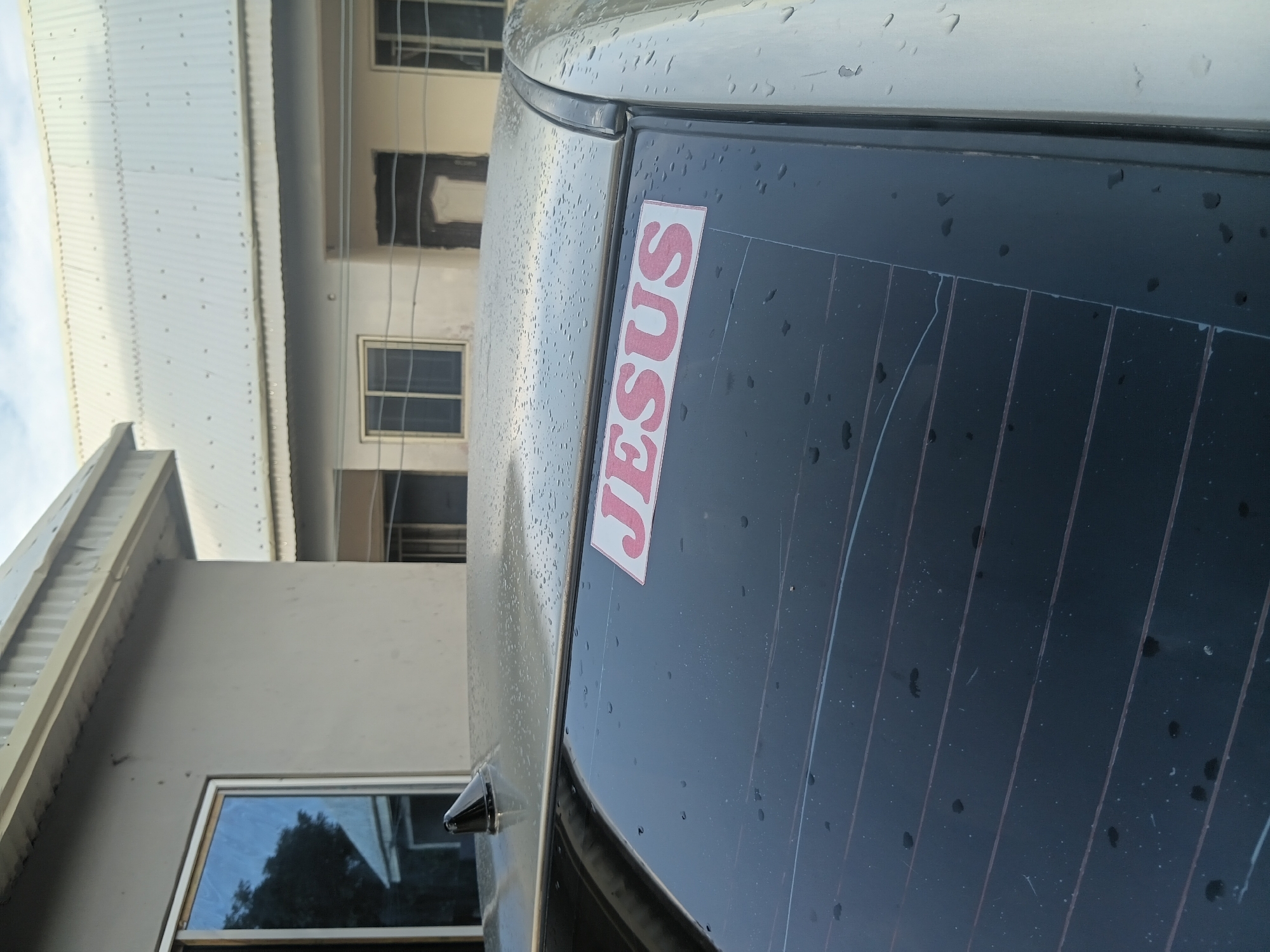 The image displays the rear section of a light-colored vehicle, likely a car, observed from a slightly elevated perspective in Jalingo, Nigeria. Water droplets are distributed across the car's roof and rear window, indicating recent rain or dew. A black shark fin antenna is mounted on the left side of the car's roof. On the upper right quadrant of the rear window, a rectangular white sticker with the word "JESUS" printed in red capital letters is prominently displayed. The rear window features embedded horizontal defroster lines and exhibits several faint scratch marks. In the background, two buildings are visible. The closer building on the left is light gray and plastered, featuring a white-framed window with dark, reflective glass. Further back and to the right, a second light-colored building with multiple dark-framed windows and a large, light-colored corrugated metal roof is present. Horizontal lines, possibly utility wires, are stretched across the mid-ground in front of this distant building. The sky above is overcast. No individuals are depicted in the scene.