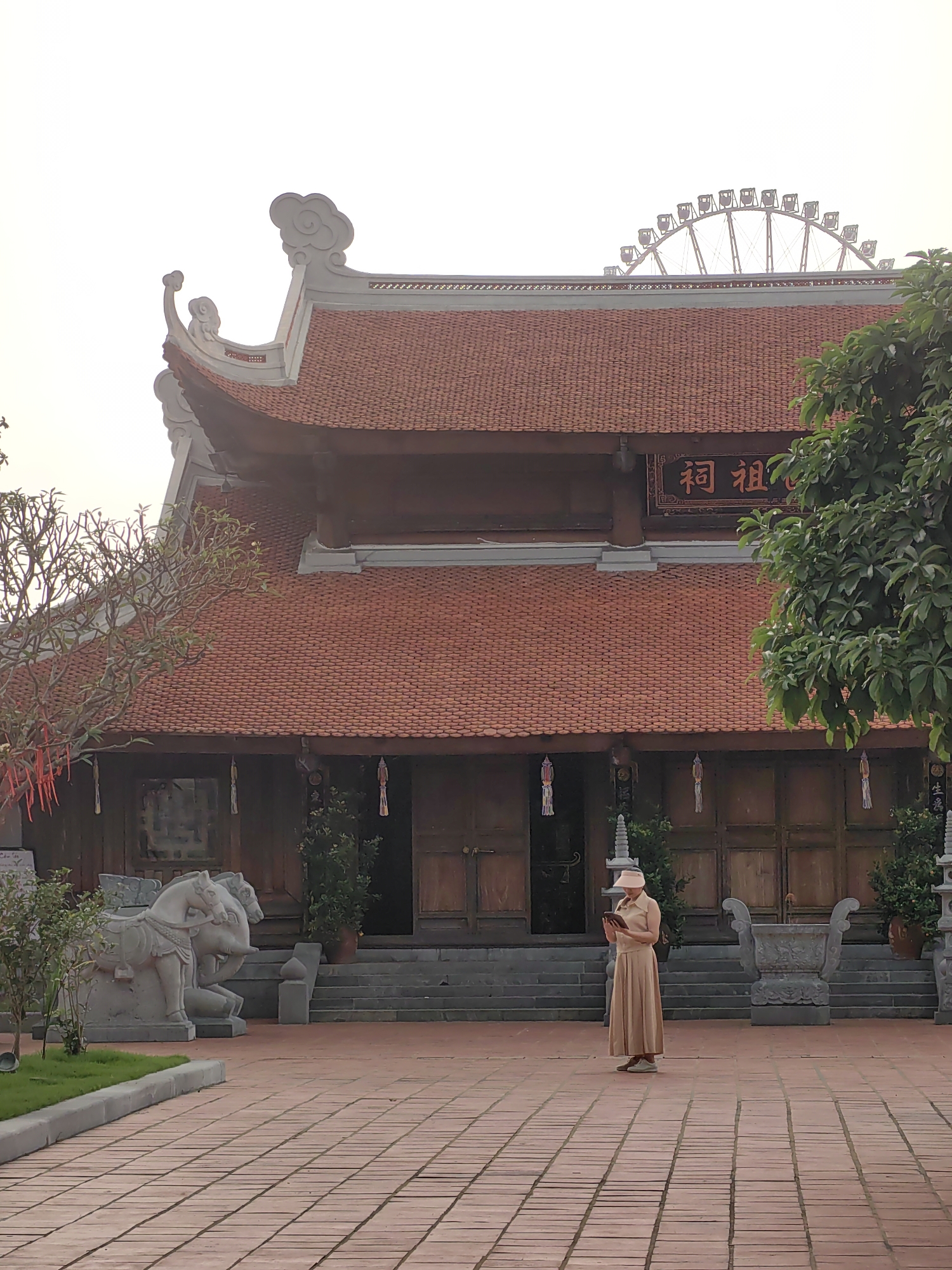 A full outdoor shot shows a woman standing on a brick-paved courtyard in front of a temple in the city of Hong Gai Ward, Vietnam. The woman faces the camera and looks down, holding a small device in her hands. She wears a tan dress, sneakers, and a pink visor. Behind her is a large, two-story temple with a red tile roof, wooden doors, and a Chinese inscription above the entrance. To the left of the temple is a stone statue of two horses, and to the right is a green tree. A Ferris wheel is visible in the distant background. The sky is overcast.