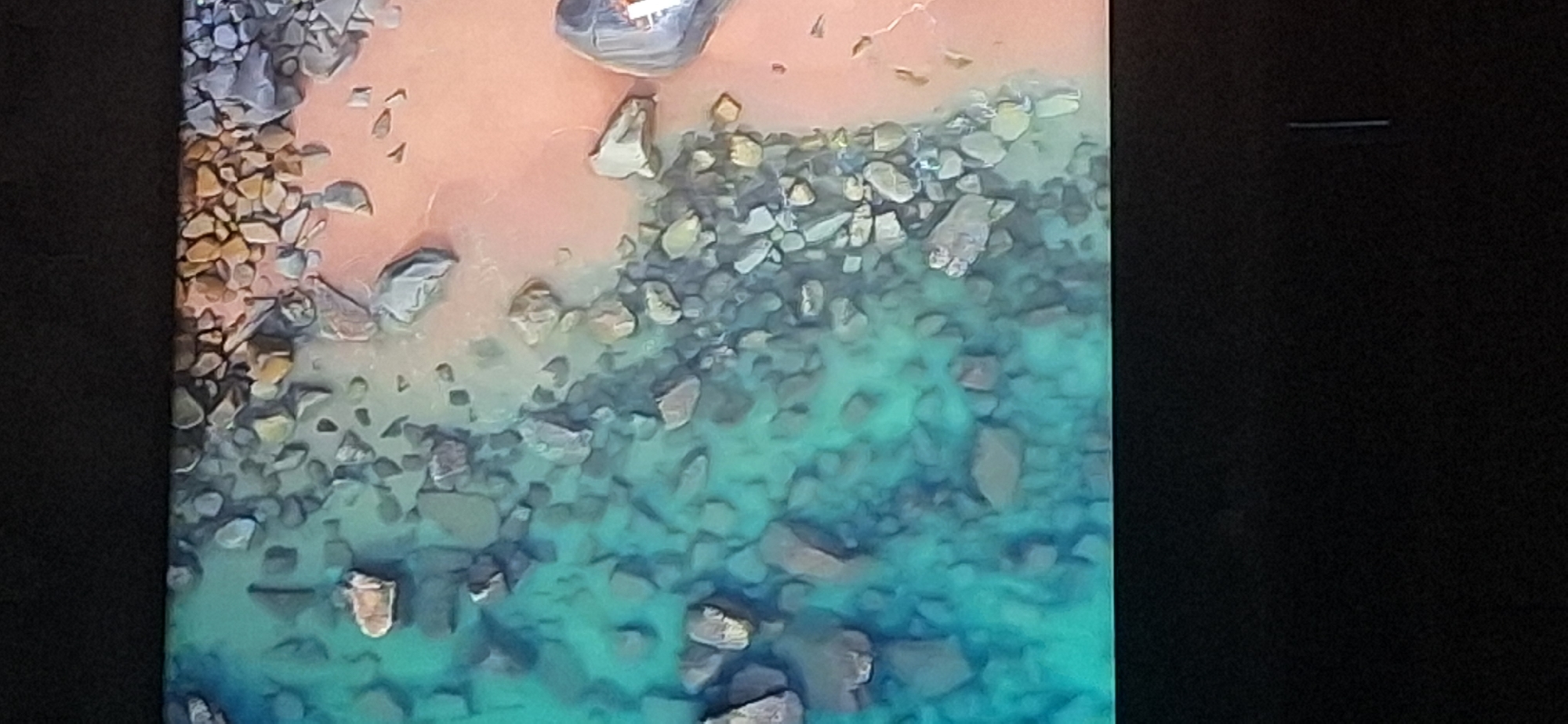 The image displays an aerial view of a rocky shoreline with clear turquoise water lapping against a sandy beach. The rocks vary in size and color, ranging from light gray to darker shades, and are scattered both in the water and on the shore. The water is shallow near the edge, allowing the submerged rocks to be visible, creating a textured pattern. The sand is a light pinkish-orange hue and appears smooth in some areas, interrupted by clusters of rocks. The overall scene is serene and natural, suggesting a coastal or lakeside environment. There are no people or man-made structures immediately visible in the frame. The image does not provide any information that would allow for the identification of its specific location within Bila Tserkva, Ukraine.