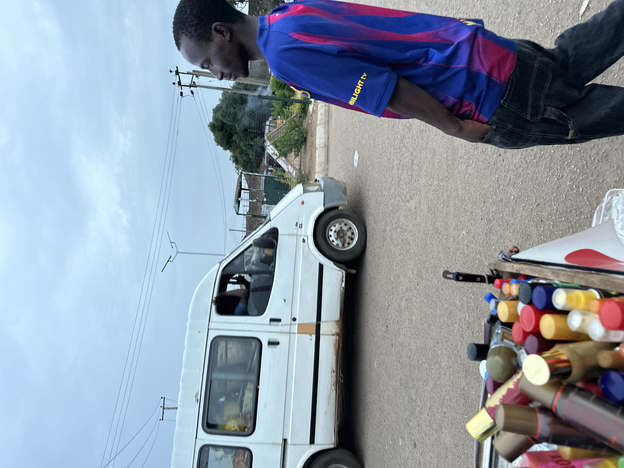 In Bukuru, Nigeria, a young Black male wearing a blue and pink striped t-shirt and dark denim pants stands on a paved road, with his right hand in his pocket. He is looking to his right. Beside him is a cart filled with various spray cans and bottles. In the background, a white minibus is driving on the road, and a utility pole with wires is visible. The sky is overcast.