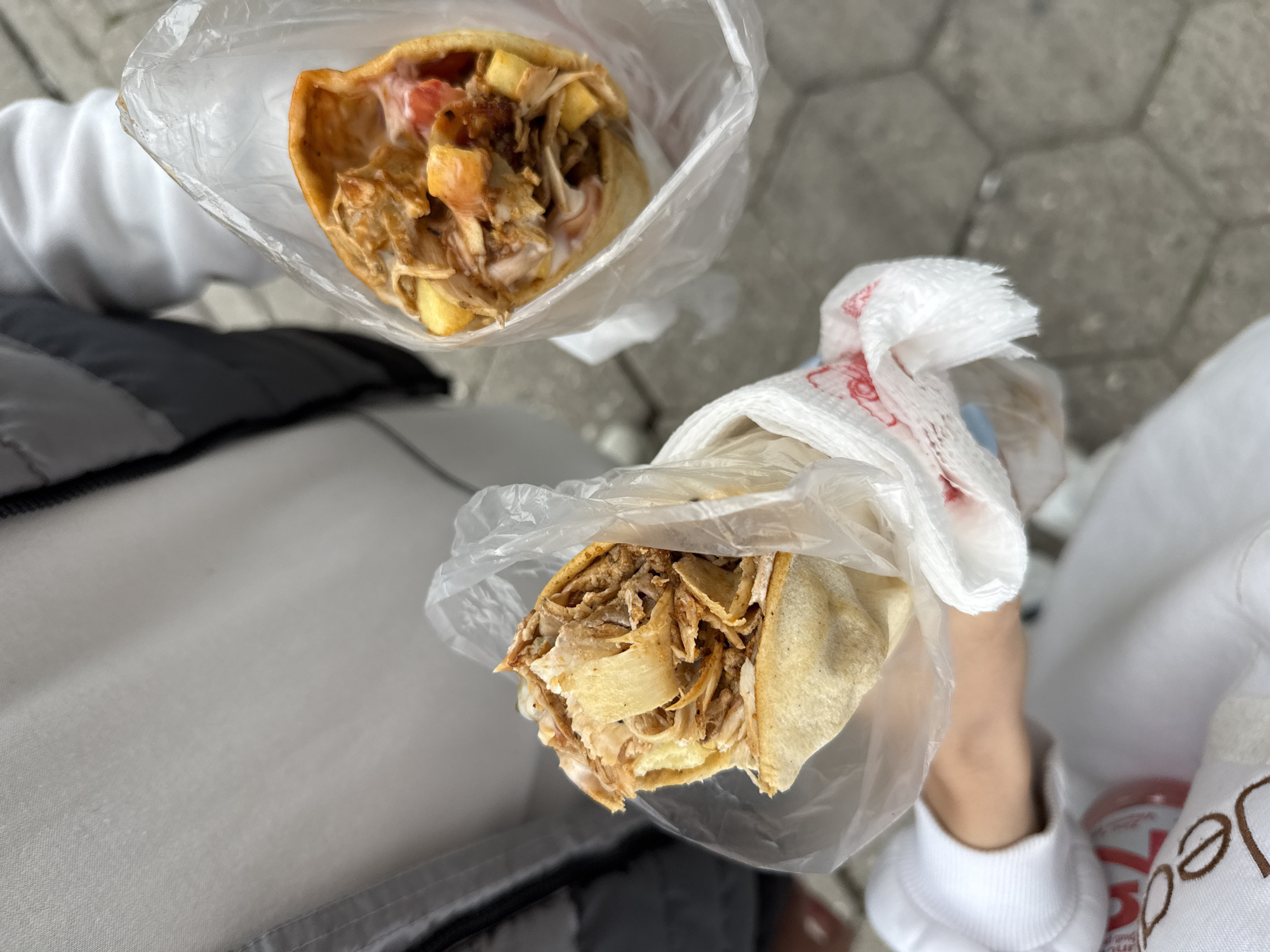 The image shows two doner kebabs held in clear plastic bags with white napkins. The kebabs contain shredded meat, sauce, and possibly vegetables; one also has fries. They are held over what appears to be a gray tiled or paved surface, and a person wearing a light-colored tracksuit is holding one. Part of a beverage can with dark lettering is visible in the lower right corner. The scene suggests an outdoor setting, and based on the information that this is in Plovdiv, Bulgaria, it is likely a food stall or street food setting.