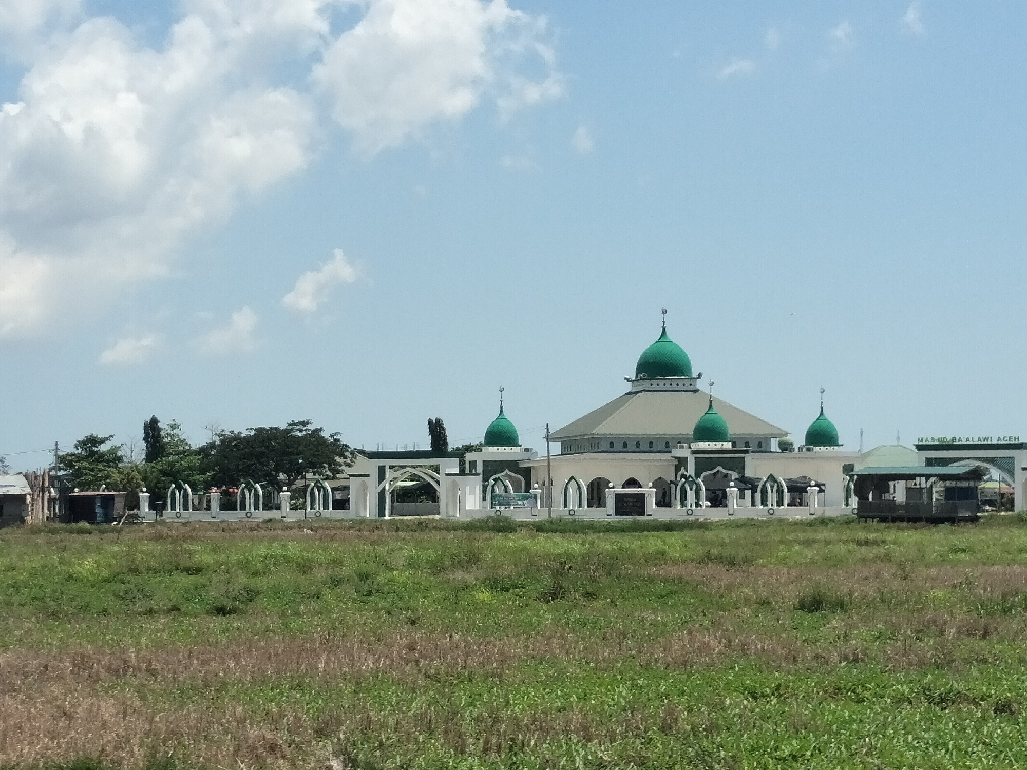 The image shows Masjid Ba'alawi Aceh, a mosque located in Matangkruet, Indonesia. The mosque features a central dome and several smaller domes, all painted green. The building is surrounded by a white fence with arched openings. In the foreground is a field of green and brown grass. The sky is blue with scattered white clouds. There are a few buildings in the distant background.