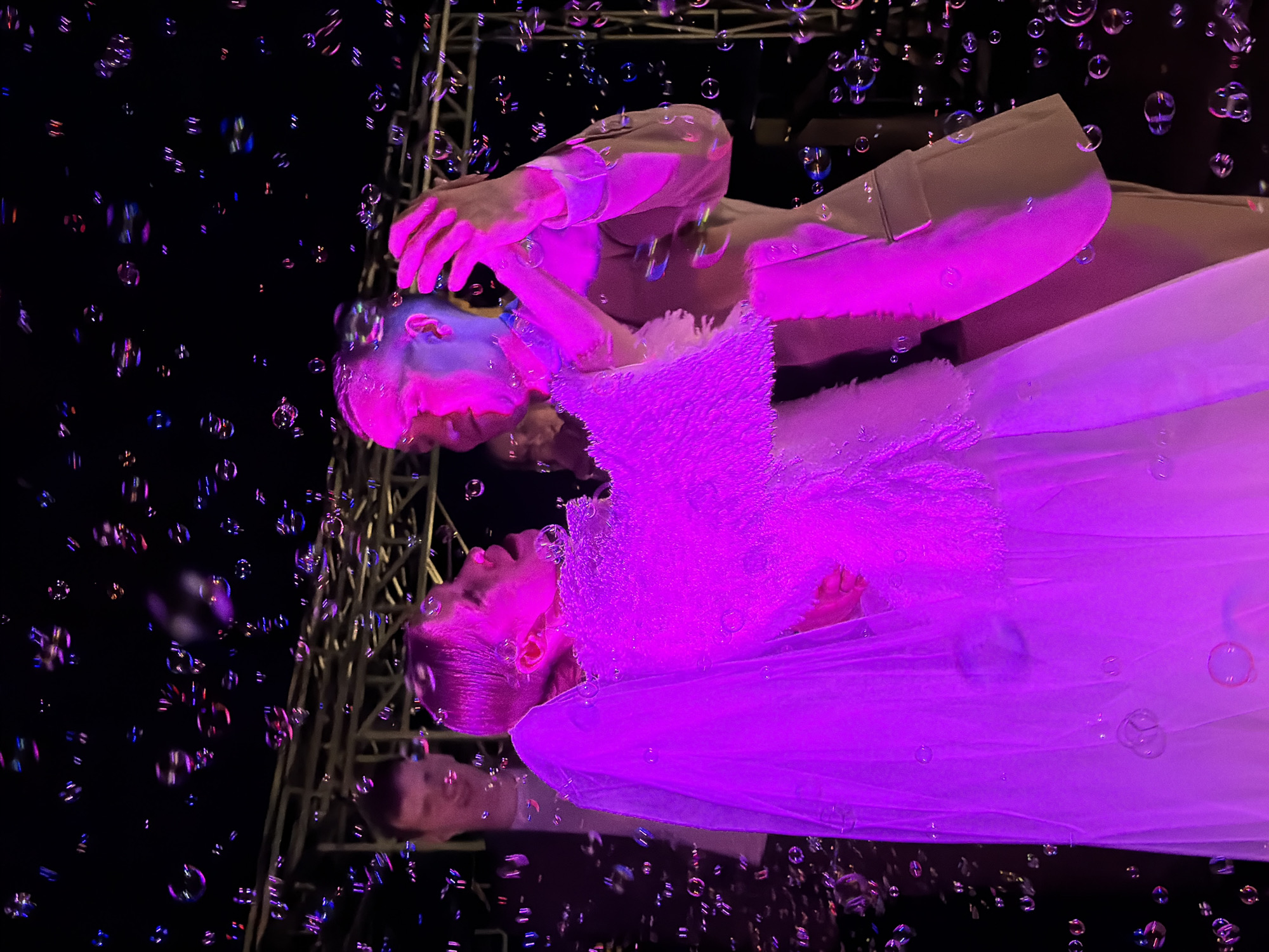 A low-angle shot captures a scene under a pink light. A woman in a white, frilly dress is looking upwards, seemingly interacting with a cascade of bubbles floating in the air. Her hands are raised near her face, as if trying to catch the bubbles. A reflection of a man, clad in a light-colored suit, is visible behind her. The background is dark, with hints of a metal structure, possibly part of an outdoor stage or setup. The setting appears to be outdoors at night, evidenced by the dark sky peppered with bubbles. This event seems to be taking place in Chaikovsky, Russia.