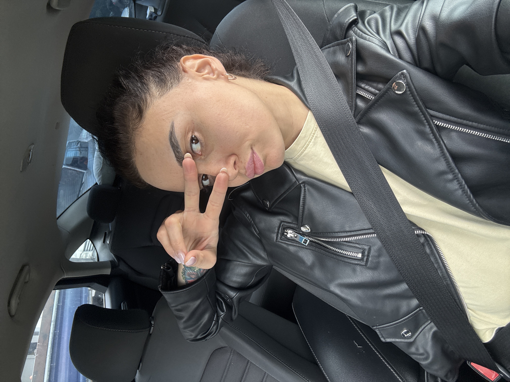 A young adult female is inside a car, wearing a black leather jacket over a light-colored shirt, and a seatbelt. She is making a peace sign with her right hand. The interior of the car is primarily black. The environment outside the car suggests an urban area, possibly the city of Tbilisi, Georgia, based on the architectural style visible through the window and the presence of a European-style building.