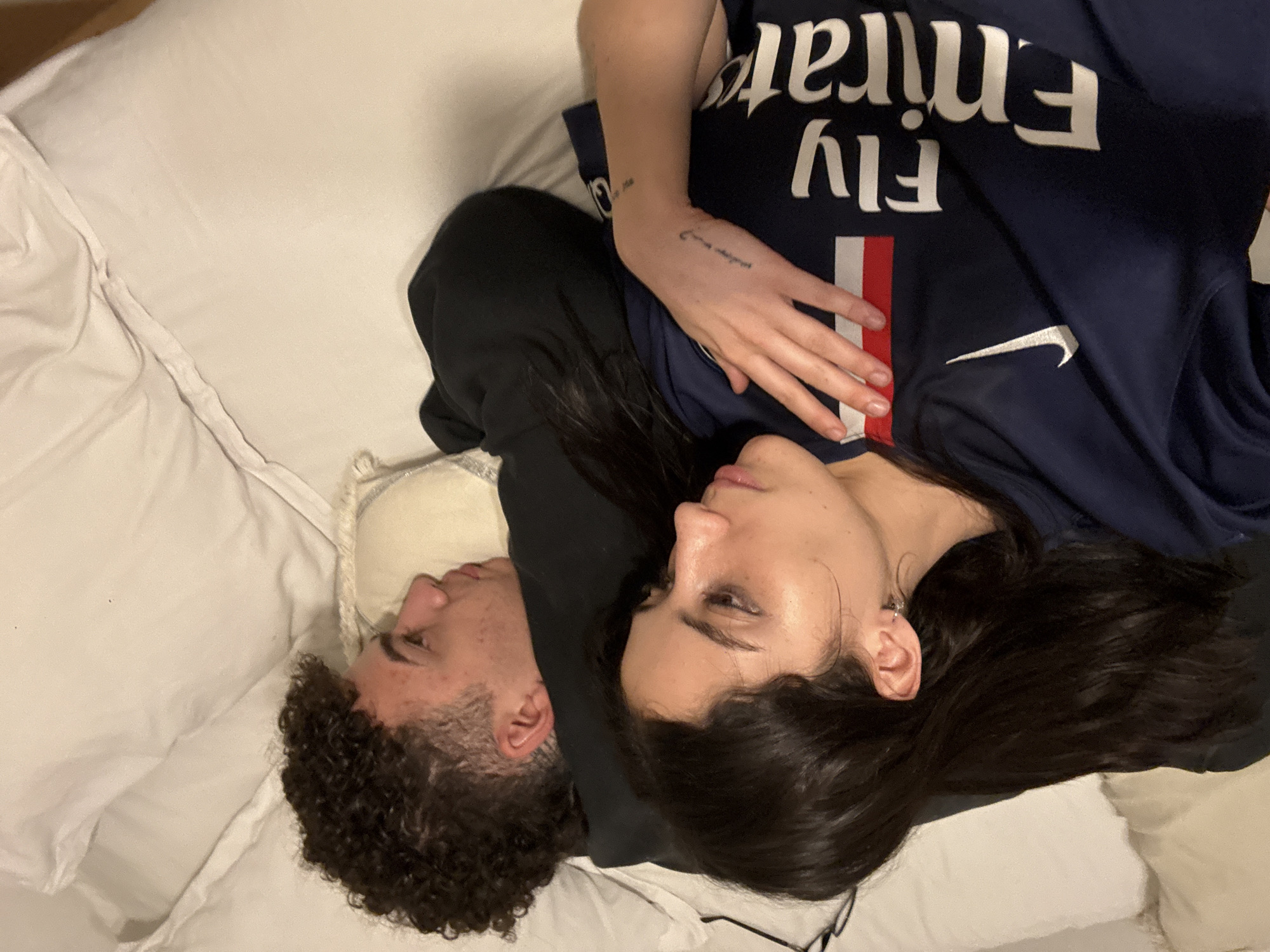 An overhead view depicts two individuals, a man and a woman, lying close together on what appears to be a bed or soft surface covered with white bedding. Their heads are positioned towards the lower right of the frame, with the man's profile and curly dark hair visible on the left, and the woman's profile and long dark hair on the right.
The woman is wearing a dark blue short-sleeved jersey, identifiable as a Paris Saint-Germain (PSG) football club shirt, featuring a red and white vertical stripe down the left side and a white Nike swoosh logo. The text "fly emirates" is visible in white letters across the chest, appearing inverted in the image. She has small script tattoos on her left forearm, one of which appears to read "tu me dis". Black-framed glasses rest near her head.
The man has dark, curly hair and his head is resting on a light-colored cushion or pillow. He is wearing a dark-colored garment, possibly a hoodie, underneath the woman's arm. Both individuals have calm, relaxed expressions, suggesting a moment of rest or intimacy. The indoor setting features soft, warm lighting, hinting at evening or nighttime. The scene takes place in Buenos Aires, Argentina.