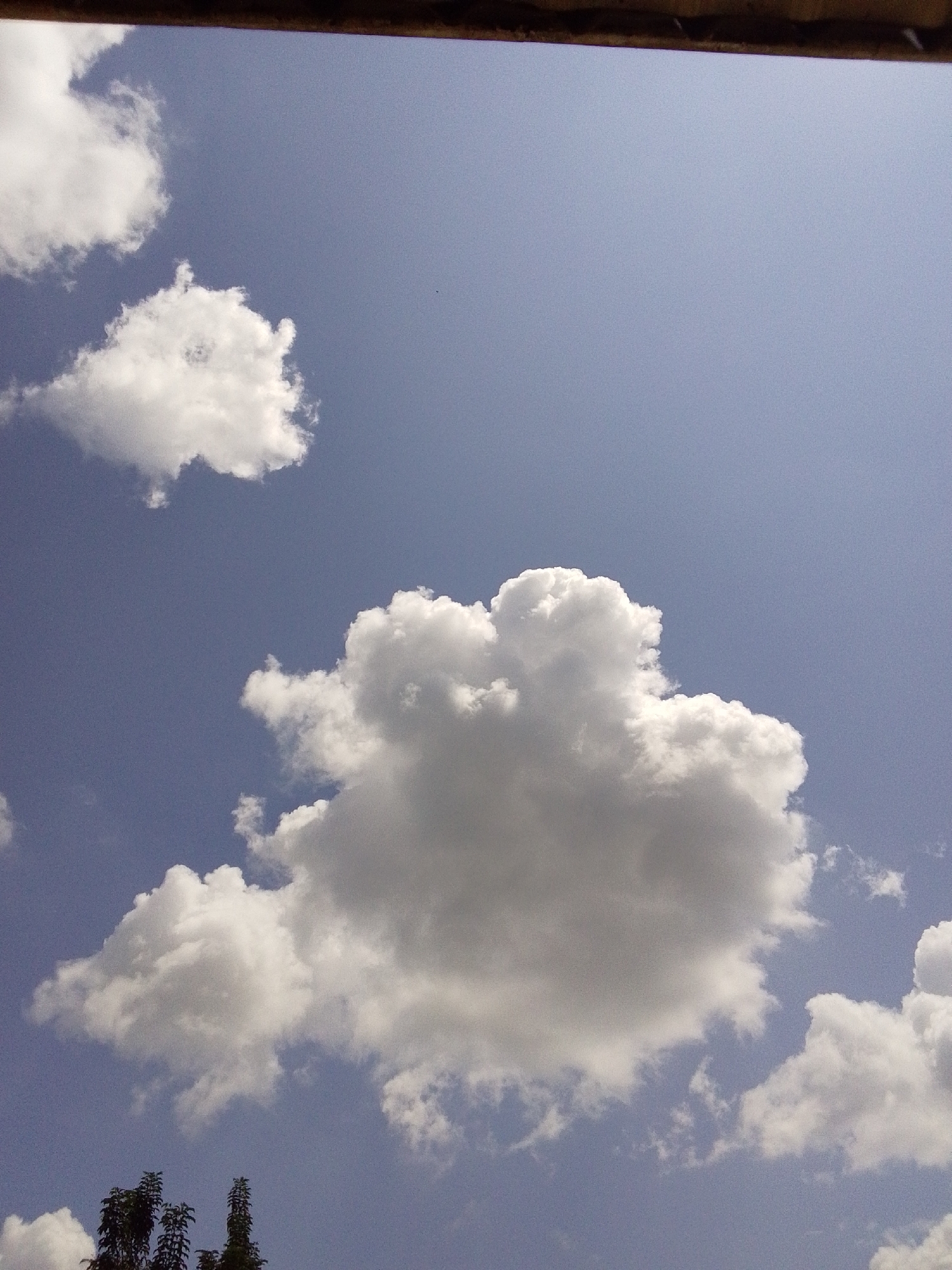 This is a photograph of the sky with clouds and some treetops visible. The sky is a clear blue. There are several fluffy white clouds of varying sizes scattered across the frame. In the foreground, at the bottom of the image, the dark green tops of trees are visible, suggesting the photograph was taken from a low vantage point looking upwards. The top edge of the image is bordered by a dark brown element, possibly a roof or a ledge. The scene appears to be outdoors on a sunny day. The location is identified as Serti, Nigeria.