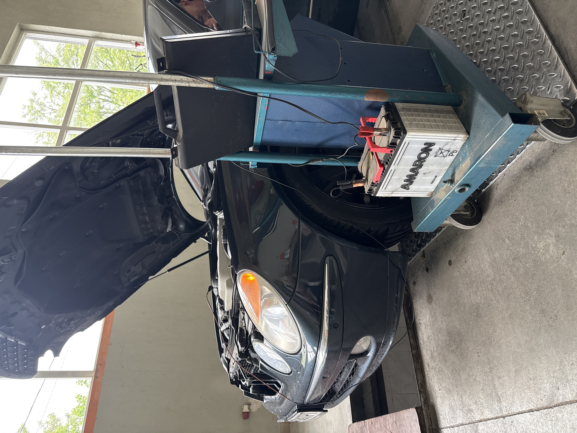 The image depicts a dark-colored car in an auto repair shop, with its hood open. A battery charger is connected to the car's battery, which is sitting on a metal stand with wheels. A person's head is barely visible behind the metal stand. The location is likely in Stara Zagora, Bulgaria, as the license plate on the car might be from Bulgaria. The environment is an indoor garage with a window in the background. The garage floor is made of concrete.