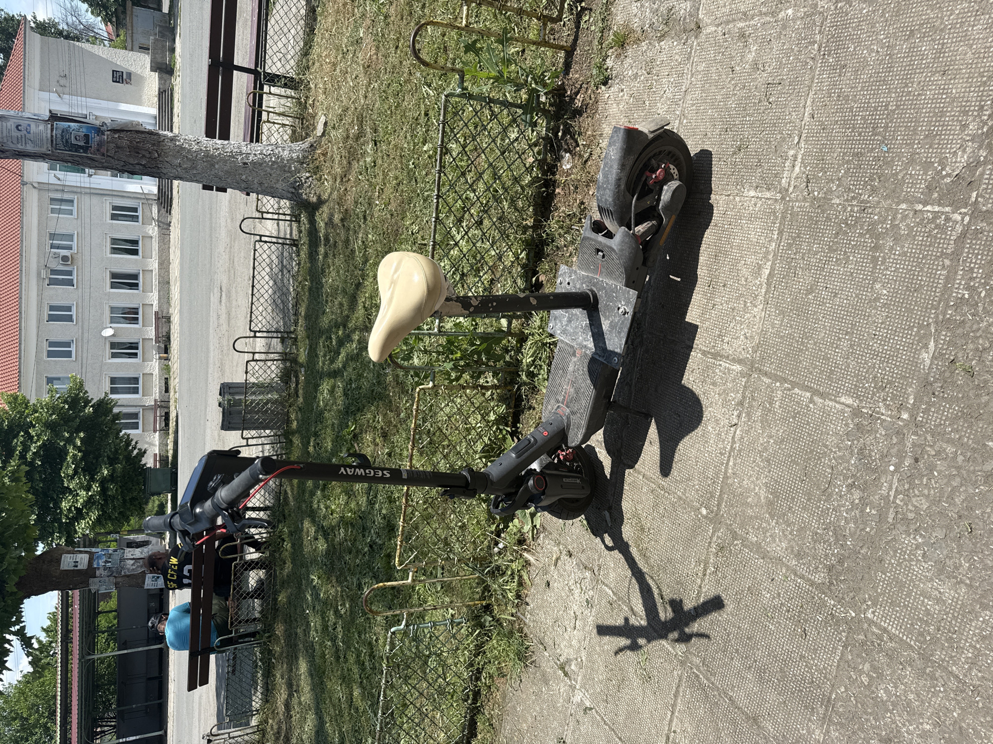 The image depicts an outdoor scene in Rusokastro, Bulgaria, on a bright, sunny day, evidenced by the strong shadows cast by objects. A black electric scooter, branded "SEGWAY" on its stem, is parked upright on a textured concrete pavement in the foreground. This scooter features a custom-fitted beige seat and red accents on its wheels/brakes. It is positioned next to a rusty metal fence and U-shaped bike racks, separating the paved area from a grassy strip with some weeds.
To the left, a large tree with a light-colored, rough bark stands tall. Its trunk has several posters or notices affixed to it, mostly unreadable, though some appear to be memorial notices with photos. Beneath the tree, two individuals are seated on a wooden bench. One person wears a light blue t-shirt and a helmet, while the other is dressed in a dark t-shirt with the visible text "GF CREW" and a black face mask. They appear to be resting.
In the background, behind the tree and fence, is a light-colored building with a red tiled roof and multiple rectangular windows across its facade. Beyond the fence and grassy area, there's a paved road or open space. The overall setting suggests a public area, possibly a park or town square.