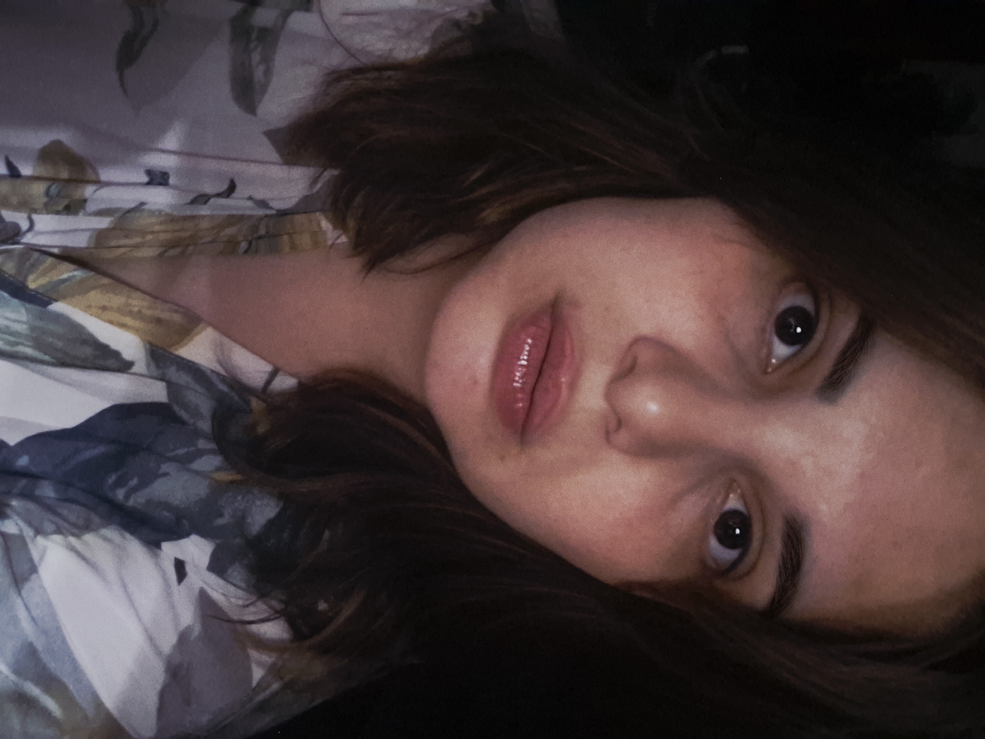 A close-up, slightly angled portrait shows a young woman with brown hair and light skin. She lies on her back, her face turned towards the camera. Her dark eyes are wide open, and her lips are slightly parted. The background includes a floral patterned fabric suggesting bedding or clothing. There is no indication of the location being Амзя, Russia.