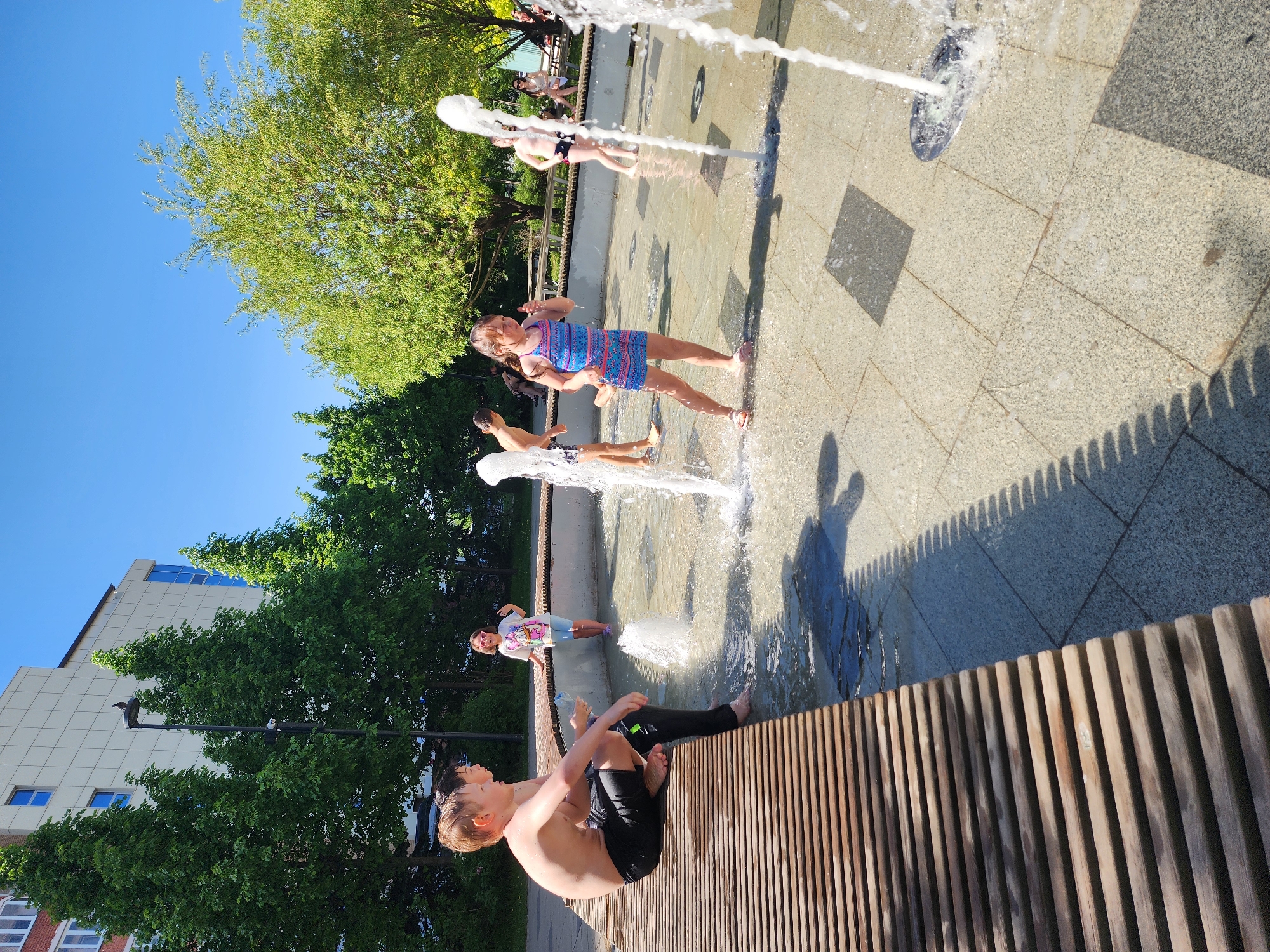 Here's a detailed description of the image:
**Location:** The image appears to be taken at a public water fountain in an urban setting. There are buildings visible in the background.
**People:**
* A group of children are playing in the water fountain. They are dressed in various summer clothes (swimsuits, dresses, shorts, t-shirts).
* At least one person is sitting on a wooden bench in the foreground.
**Subjects/Objects:**
* The central focus is the water fountain with jets of water spraying upwards.
* Wooden bench in the foreground.
* Trees and some building.
* A building is visible in the background.
* Street lamp post.
**Actions/Interactions:**
* Children are playing in the fountain, interacting with the water.
* At least one person is sitting on the wooden bench.
**Environment:**
* Sunny, bright day.
* The fountain is surrounded by a paved area.
* Trees provide some shade in the background.