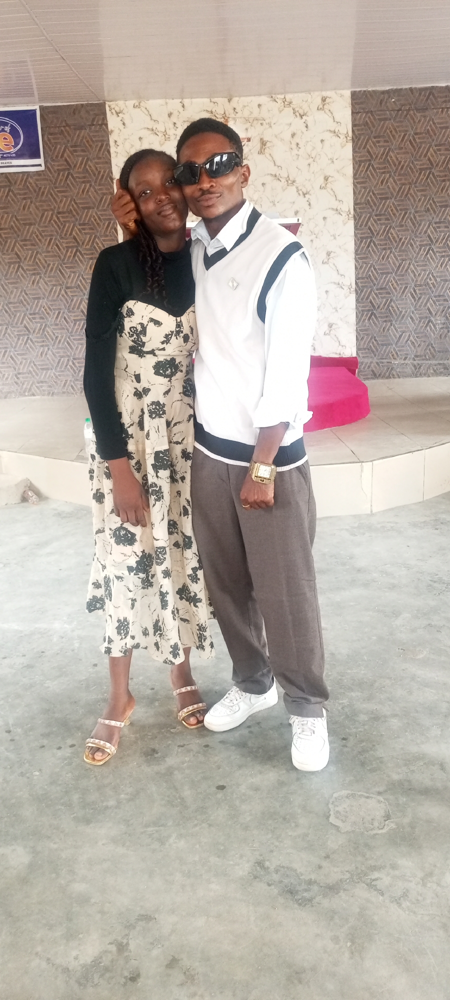 Two individuals are standing closely together in what appears to be an indoor space. The person on the left is wearing a floral-patterned dress with a black top and gold sandals, and they have their arm around the person on the right. The person on the right is wearing sunglasses, a white sweater vest over a collared shirt, grey pants, and white sneakers. A gold watch is visible on their wrist. The background includes a patterned wall, a raised platform with a pink cushion, and what appears to be a concrete floor. A sign is visible in the top left corner. The location is Lokoja, Nigeria.