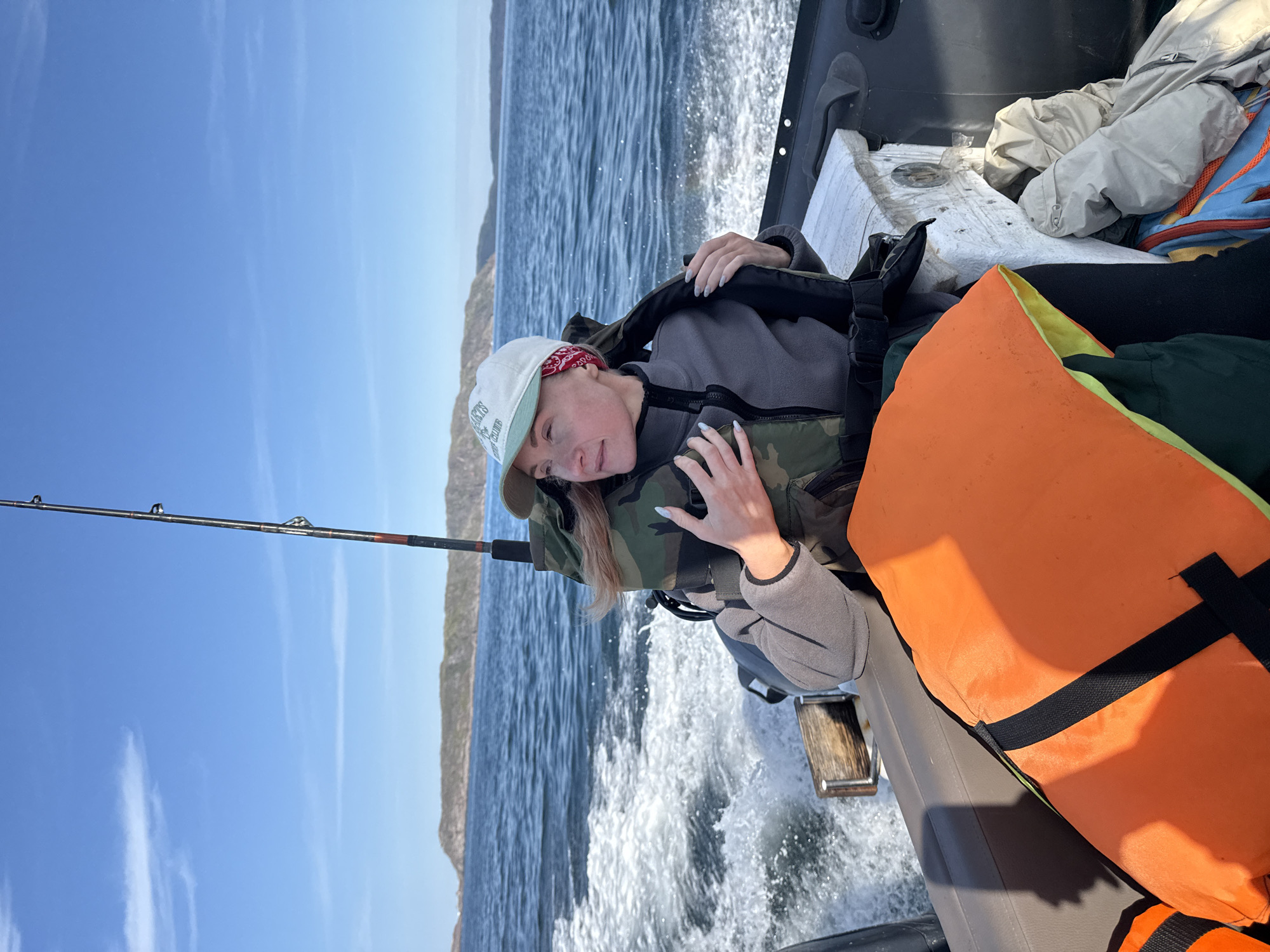 A woman wearing a camouflage life vest, grey jacket, and a white baseball cap with a red bandana around her neck is seated on a boat. She is looking to her right, away from the camera, with her hands on the life vest. In the foreground, an orange life vest is visible, partially obscuring other items on the boat. The boat is moving, creating a wake in the choppy blue water. In the background, a long, hilly island or coastline is visible under a clear blue sky. The scene appears to be taking place in Teriberka, Russia, a coastal village known for its dramatic landscapes and proximity to the Arctic Ocean.