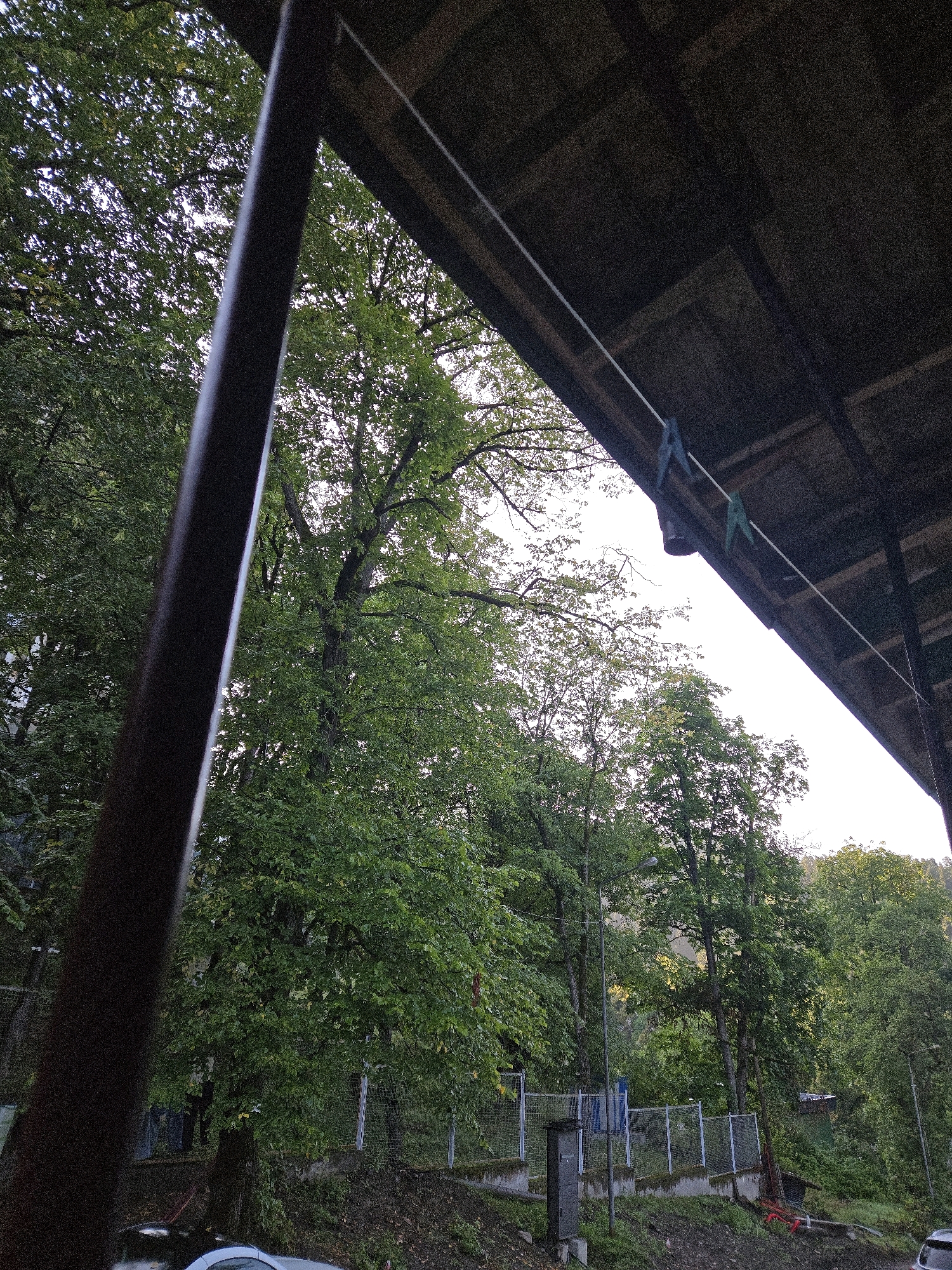 The image depicts an outdoor scene, likely a backyard or a communal area, with lush green trees dominating the background. In the foreground, a dark, cylindrical pole, possibly part of a structure or railing, is prominently positioned on the left. Above, a wooden overhang or roof is visible, with a clothesline strung across it, holding two clothespins, one blue and one green. The sky is bright, suggesting daytime.
The middle ground reveals a wire fence bordering an area with some utility boxes or structures. Beyond the fence, the dense foliage of trees continues, with some hints of a hilly landscape in the distance. The ground in the foreground appears to be dirt and debris, with a car visible at the bottom left corner.
The scene is devoid of any people. The overall impression is one of a natural, somewhat rustic setting, possibly in a residential area. The presence of the clothesline suggests everyday life and domestic activities. The bright sky and verdant trees indicate a possibly temperate climate.
Considering the details and typical landscapes of Abastumani, Georgia, this scene could be located in a residential area or a park within the town, where private homes or guesthouses often have similar architectural elements like wooden overhangs and clotheslines, set against the backdrop of the town's characteristic lush, mountainous terrain.