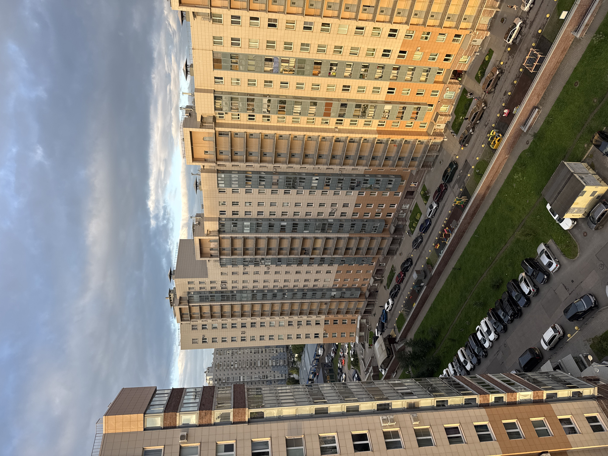 The image shows a view from above, looking down at a residential area with several high-rise apartment buildings. The buildings are modern, with light-colored facades and balconies. There is a street with cars parked along one side and a green grassy area with a playground. The sky is cloudy with hints of blue peeking through. The scene is likely in the late afternoon or early evening, as indicated by the warm lighting on the buildings. The environment appears to be urban, with multiple residential buildings close together. There are a few people visible walking on the street and near the playground. The overall impression is one of a contemporary residential development.