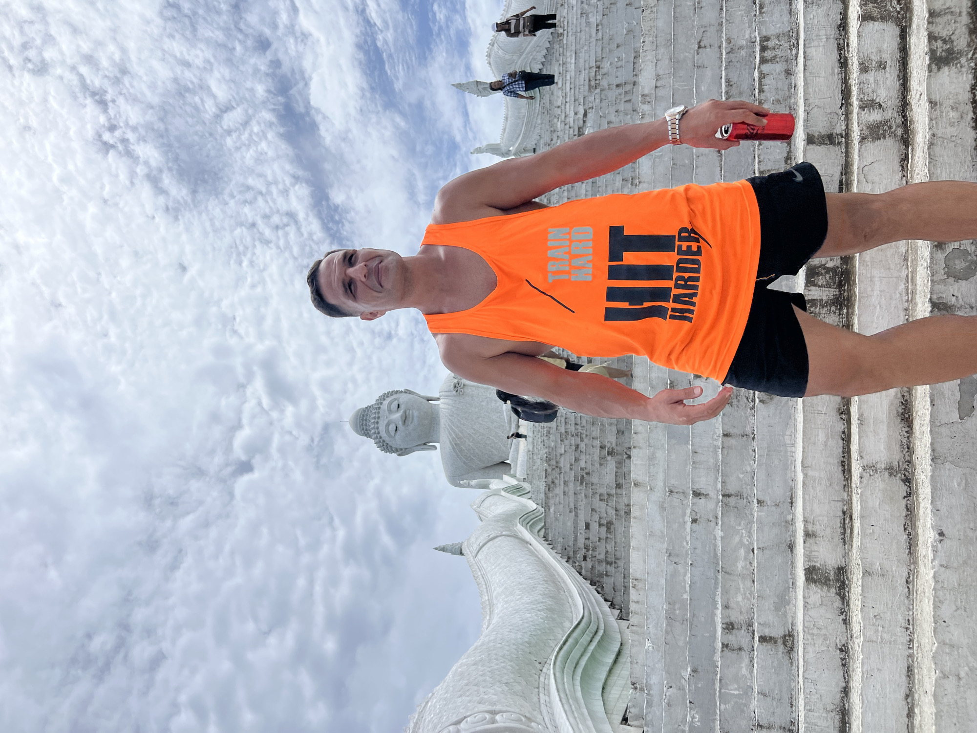 The image depicts a man standing on a wide, white concrete staircase, possibly leading to a religious site. He is wearing an orange tank top with the text "TRAIN HARD HIT HARDER" in black letters, and black shorts. On his left wrist, he wears a silver watch. In his right hand, he holds a red object, possibly a phone or a can.
In the background, a large, white statue of Buddha is visible, with a long, serpentine-like structure also made of white stone winding up the stairs in front of it. Several other people are seen further up the stairs, appearing to be tourists. The sky is cloudy, suggesting it is daytime, likely overcast or partly sunny. The overall scene appears to be outdoors, in a location with significant architectural and religious elements, consistent with a temple or monument in Karon, Thailand. The man's expression is a slight smile.