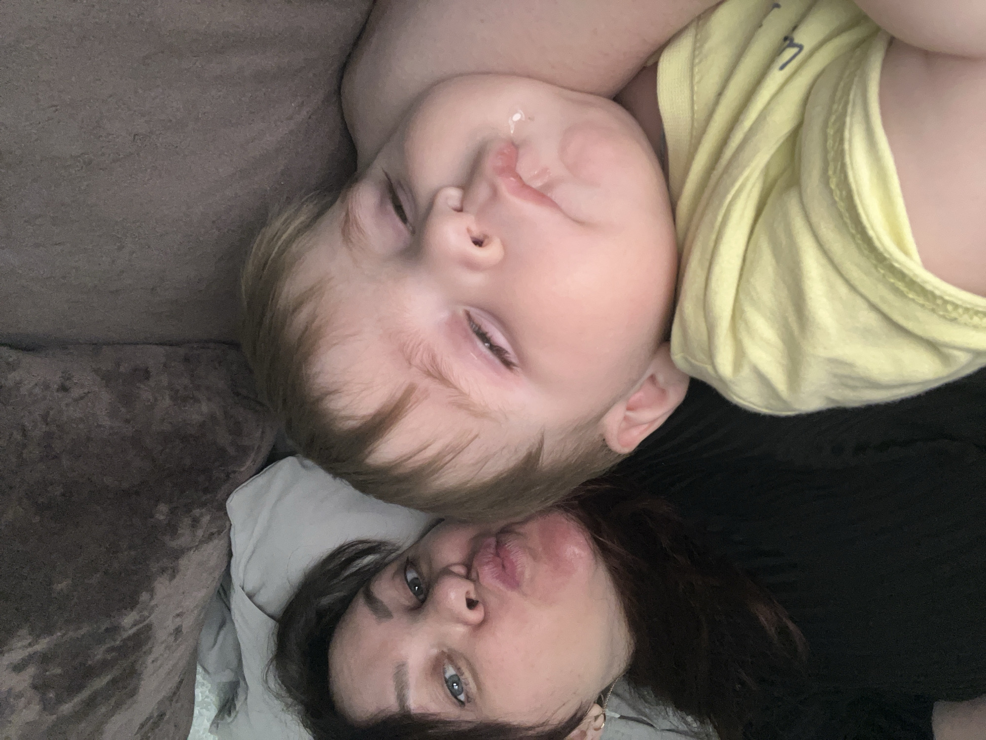 A woman and a baby are lying down together. The woman is on her back, and the baby is on their side, lying on the woman. The woman has dark hair and blue eyes, and is making a kissing face. The baby has fair hair and fair skin, and is wearing a yellow shirt. The location cannot be determined from the image.