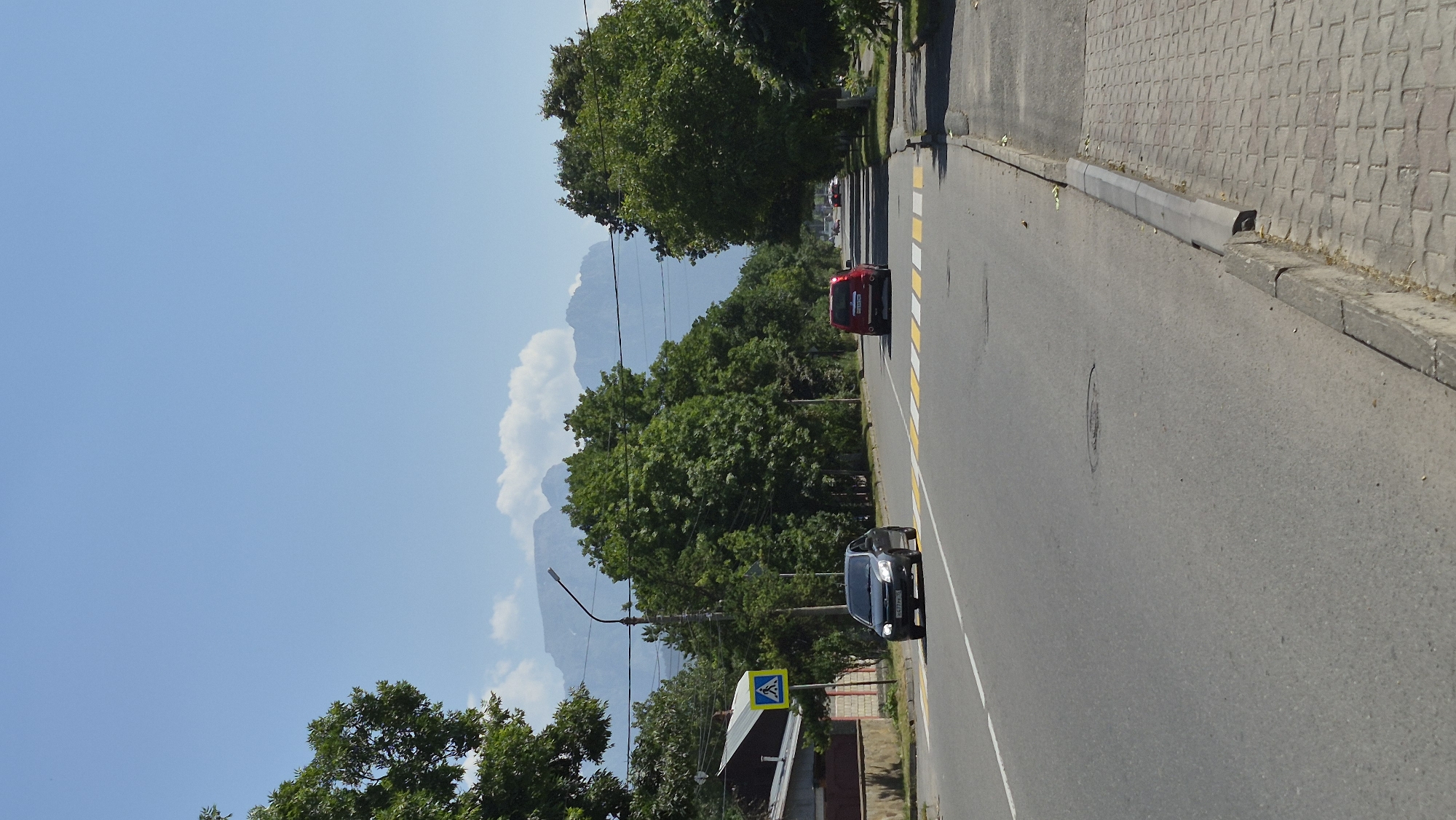 A street in Vladikavkaz, Russia, on a sunny day. The road runs towards a distant mountain range with clouds. The street is lined with green trees on both sides. A gray car is driving on the road, passing a pedestrian crossing. A red car is behind it. The sidewalk is paved with light-colored bricks. A yellow sign with a pedestrian symbol is visible on the left side of the road. There is a street lamp on the left side of the road. The sky is clear blue.