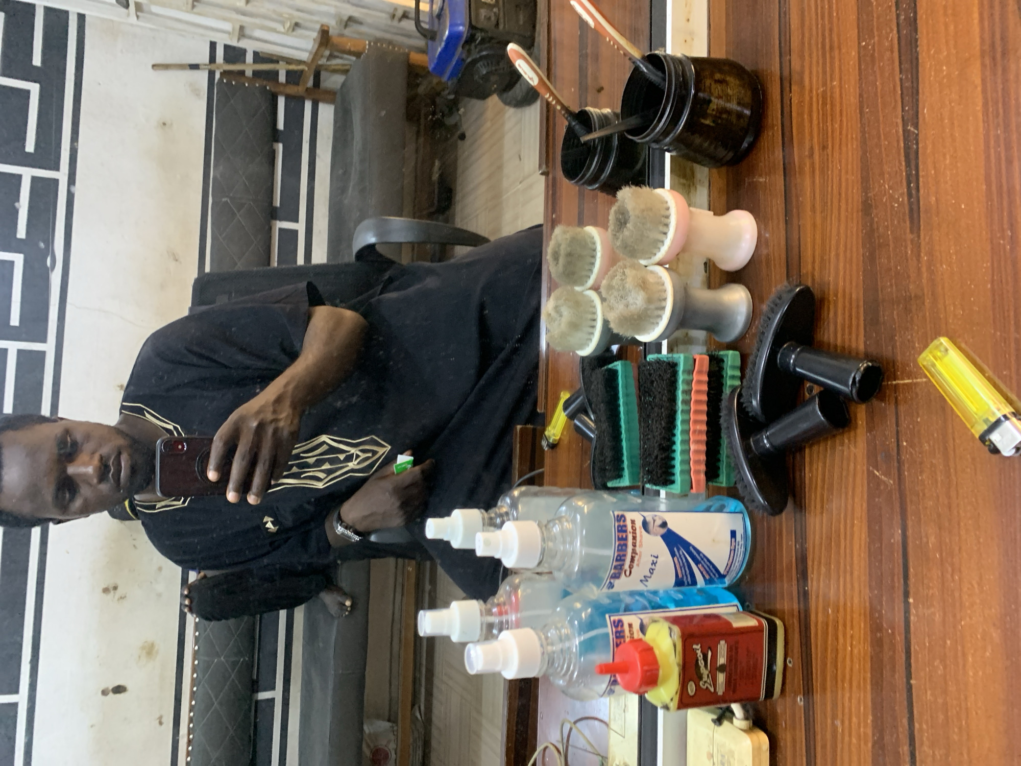 This media file shows a man in a barber shop. He is wearing a black traditional outfit with gold embroidery. He is holding a smartphone and taking a selfie in a mirror. In the reflection, we can see a barber's chair and a wall with a black and white striped pattern. On the wooden countertop, there are several items related to barbering, including brushes, spray bottles, and a lighter. The bottles appear to be hair products, with one labeled "BARBERS COMPANION Maxi". The overall impression is that of a barber at work, perhaps taking a moment to document his environment. The lighting suggests it could be daytime. The location is identified as Jos, Nigeria.