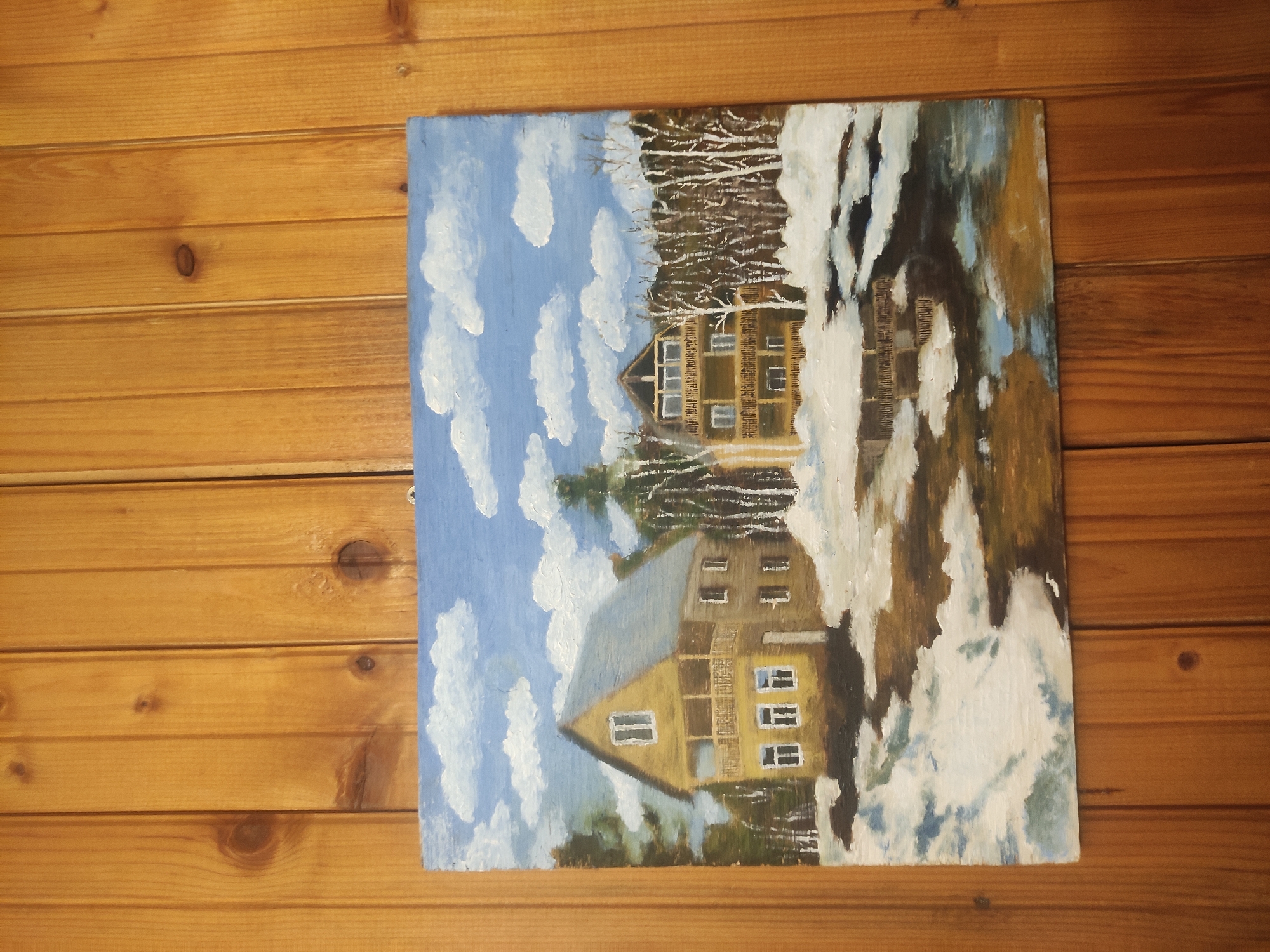 The image shows a painting of a two-story house with a gable roof, depicted in a winter landscape under a cloudy sky. The house is light brown with white window frames. A thin layer of snow covers the ground and some surrounding trees. The painting hangs on a wall made of horizontal wooden panels. The city and location are unknown.